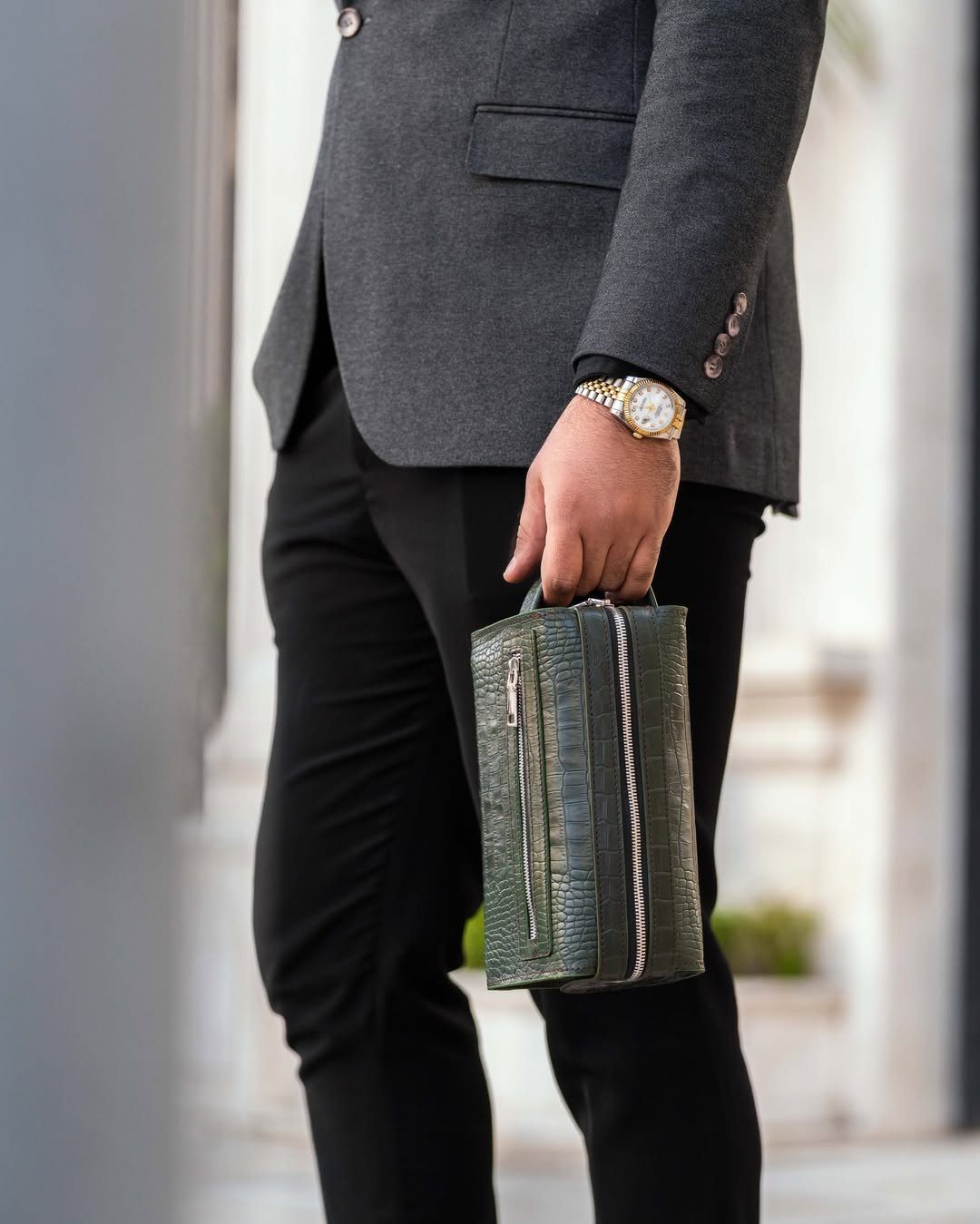 Man in a grey blazer holding a green crocodile-skin fashion clutch purse.