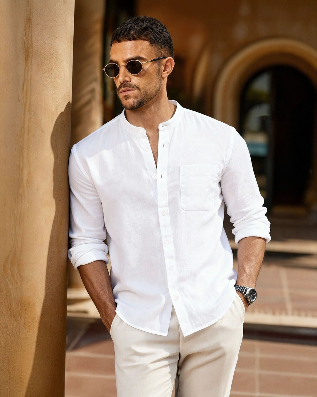 Man in white linen band collar shirt and sunglasses leaning on a pillar.