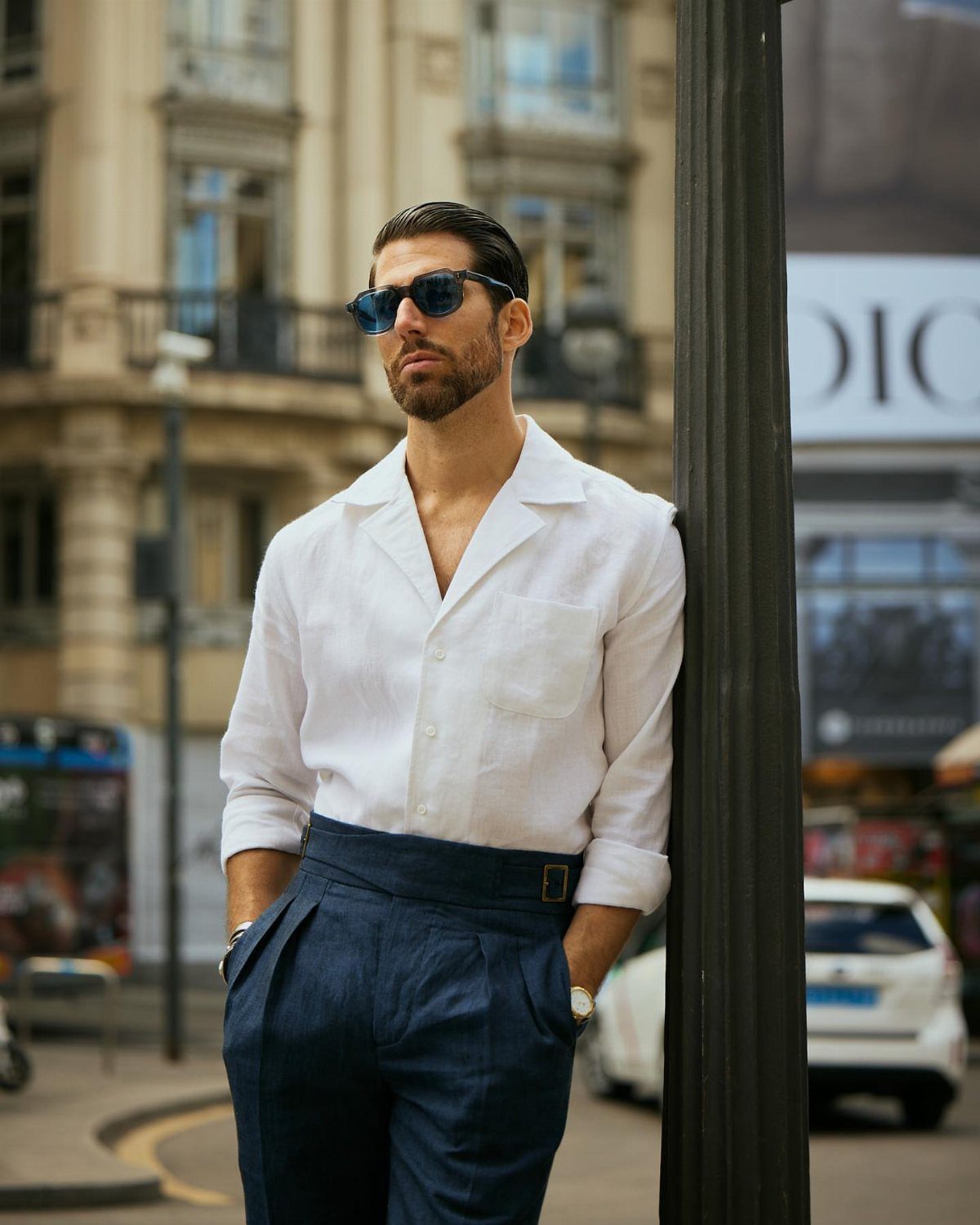 Man in white Cuban collar shirt and navy trousers leaning on a city lamppost.