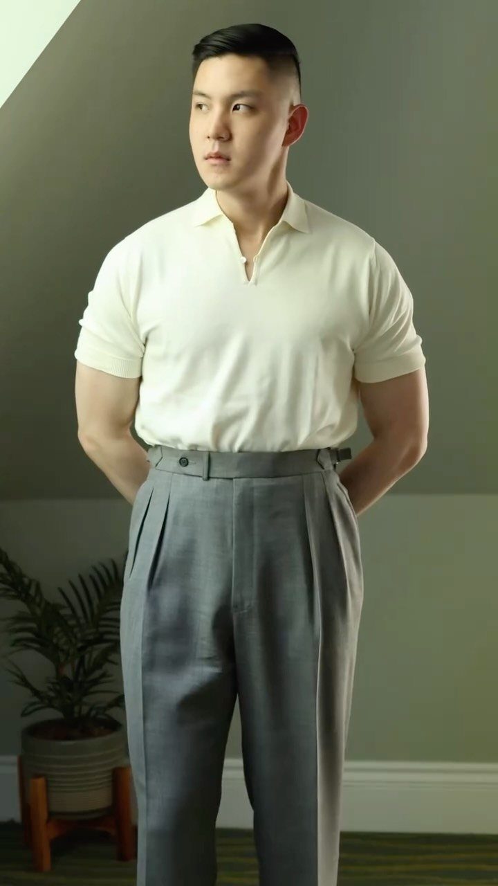 Man in a cream knit polo shirt and grey pleated trousers posing indoors.
