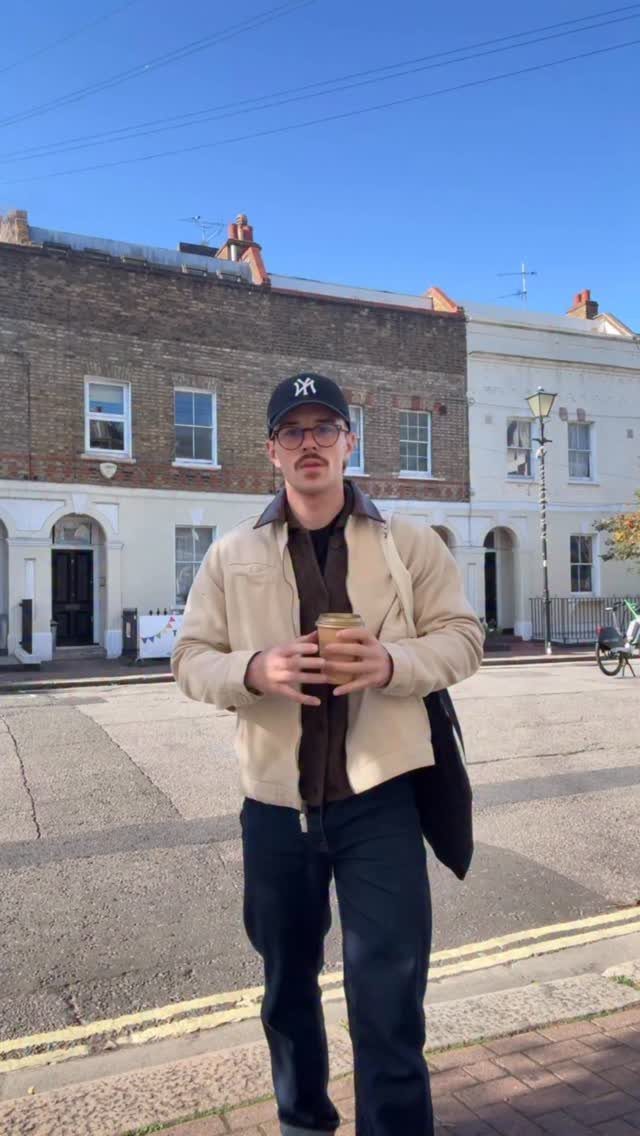 Slender man wearing a beige jacket layered over a brown shirt and dark trousers.