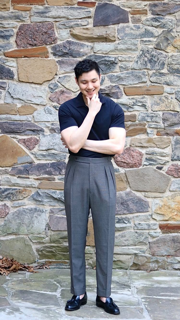 Man in black polo and grey straight-leg trousers posing against a stone wall.