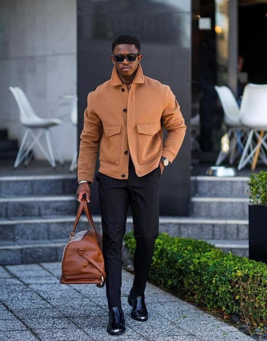 Man in a tan structured jacket and black trousers carrying a brown leather bag.