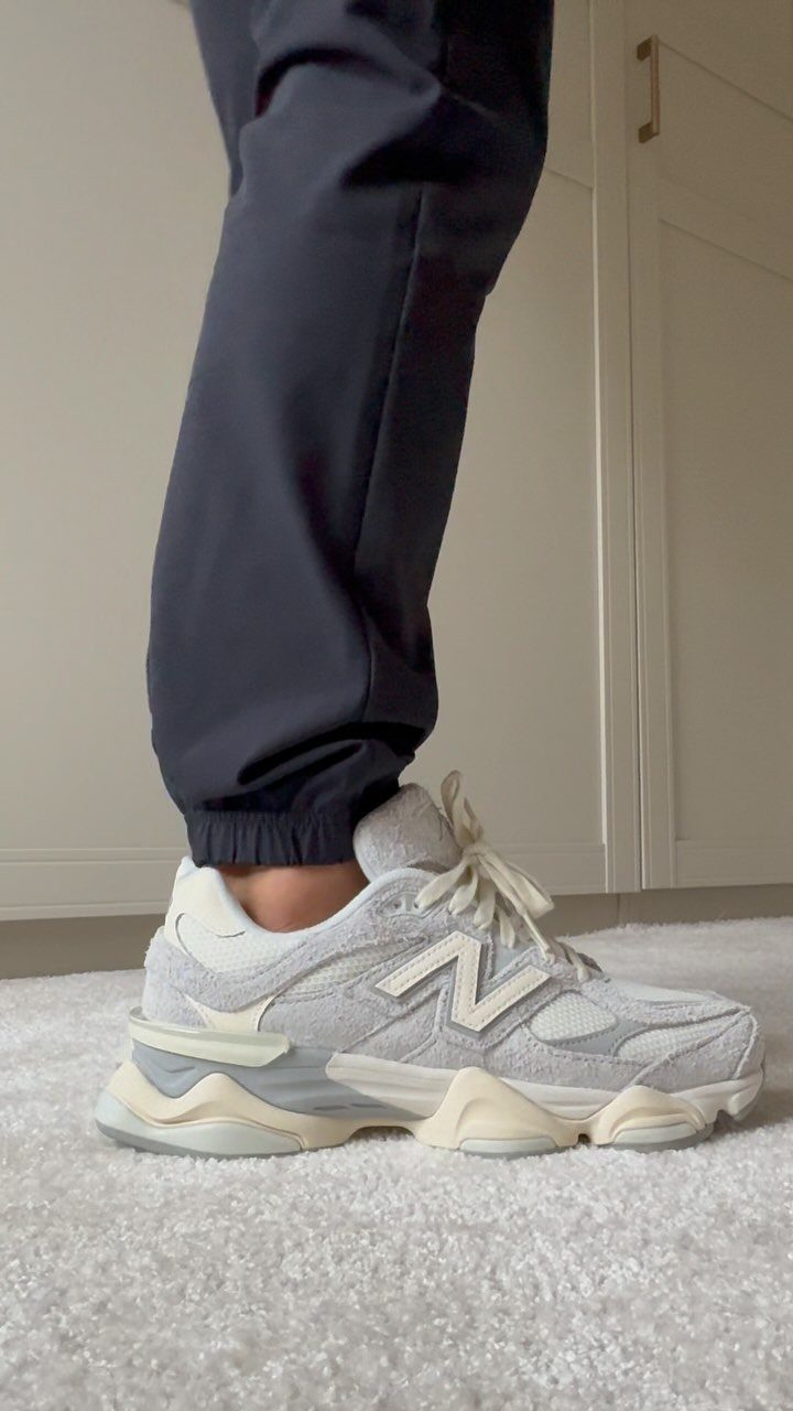 Grey and white New Balance 9060 sneakers worn with navy blue joggers on white carpet.