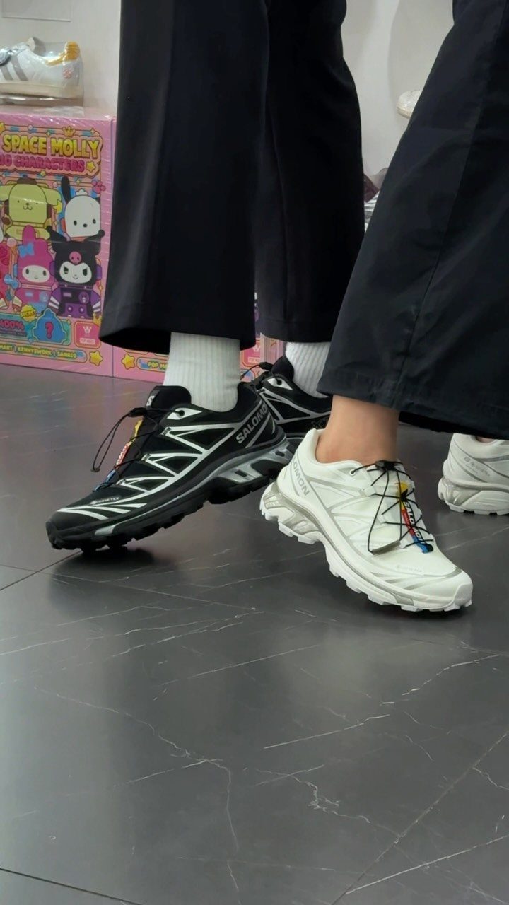 Black and white chunky Gorpcore sneakers worn with black trousers on a grey floor.