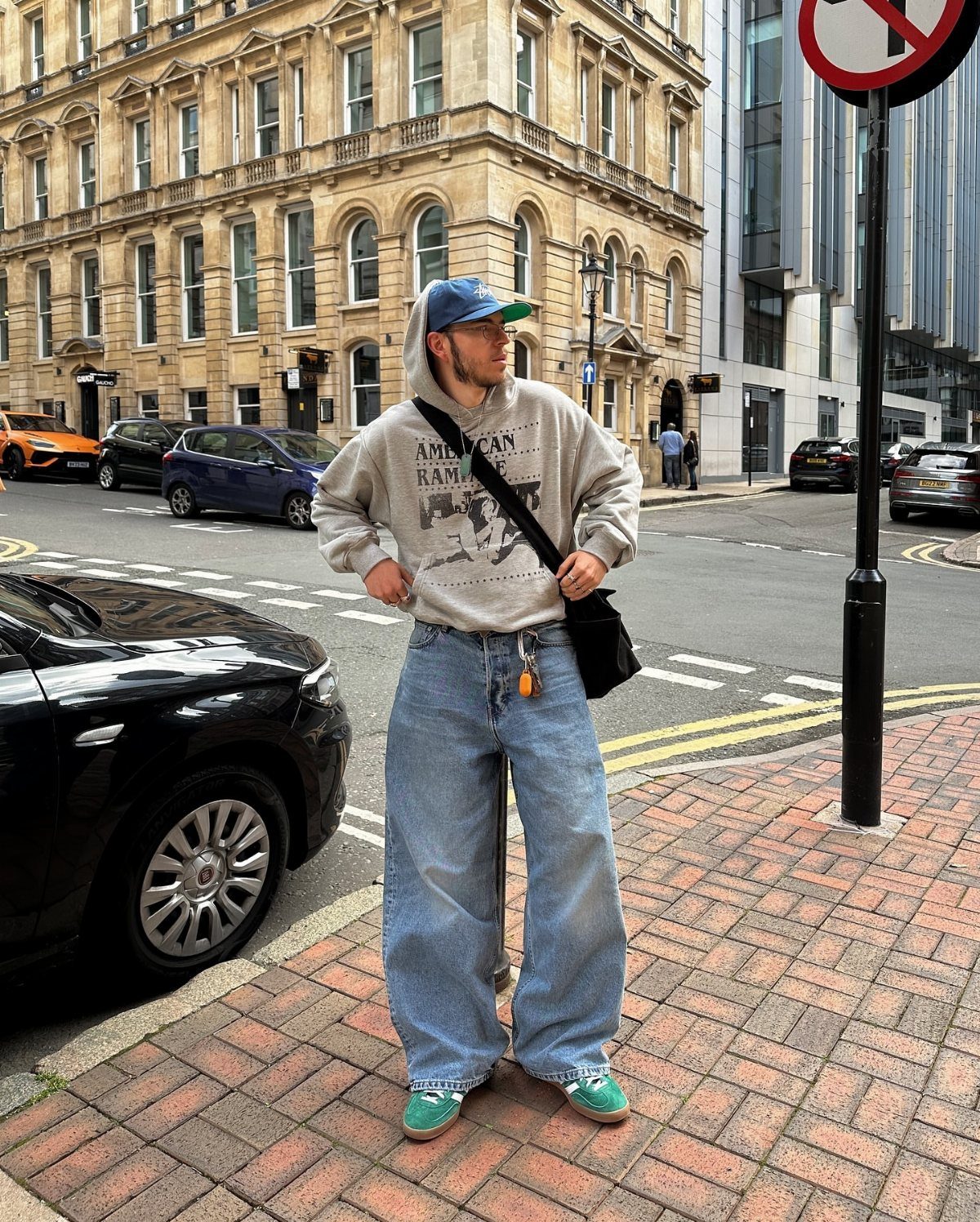 Man in wide-leg jeans and green sneakers standing on a city brick sidewalk.
