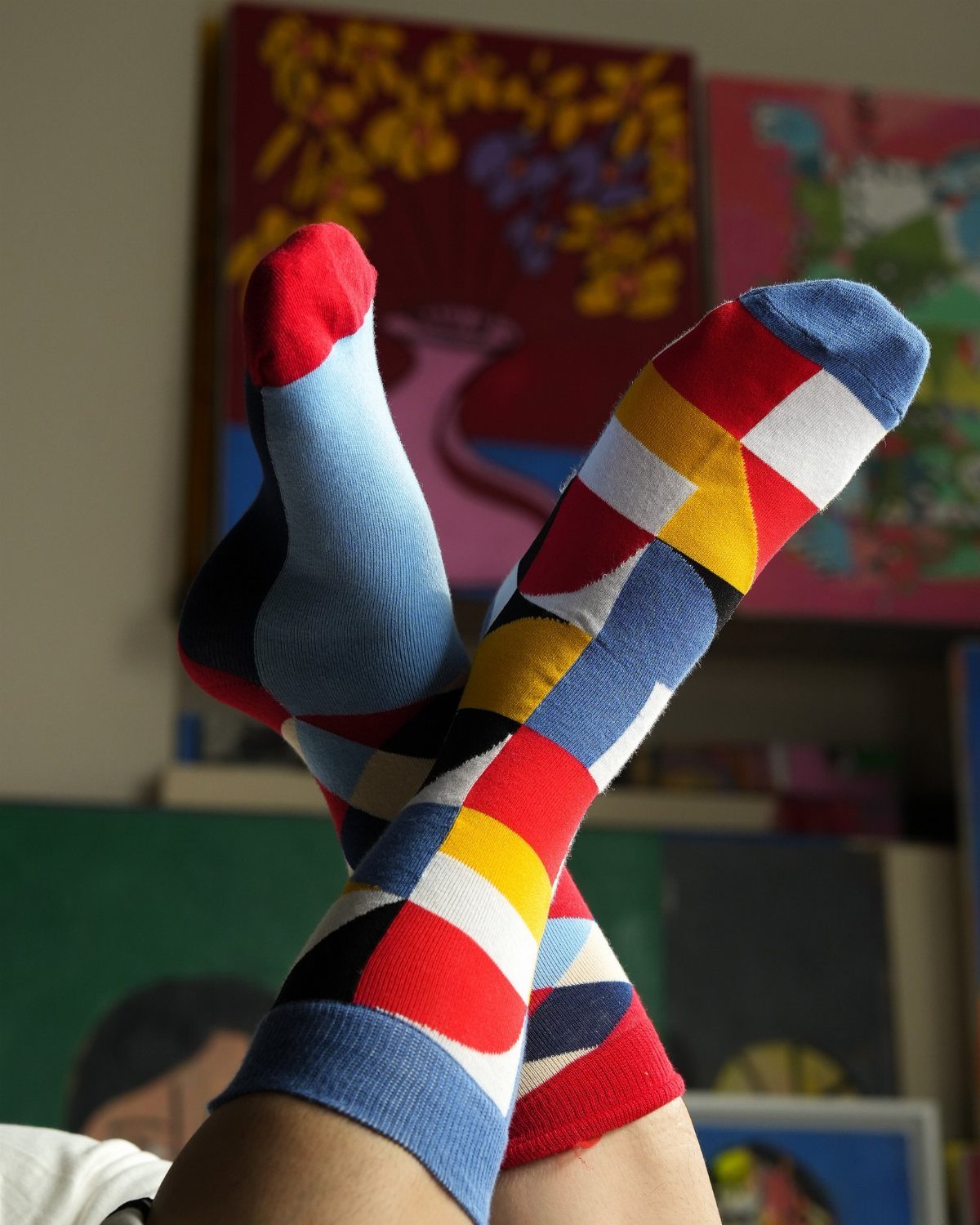 Colorful geometric patterned fashion socks worn on feet raised against a background of paintings.