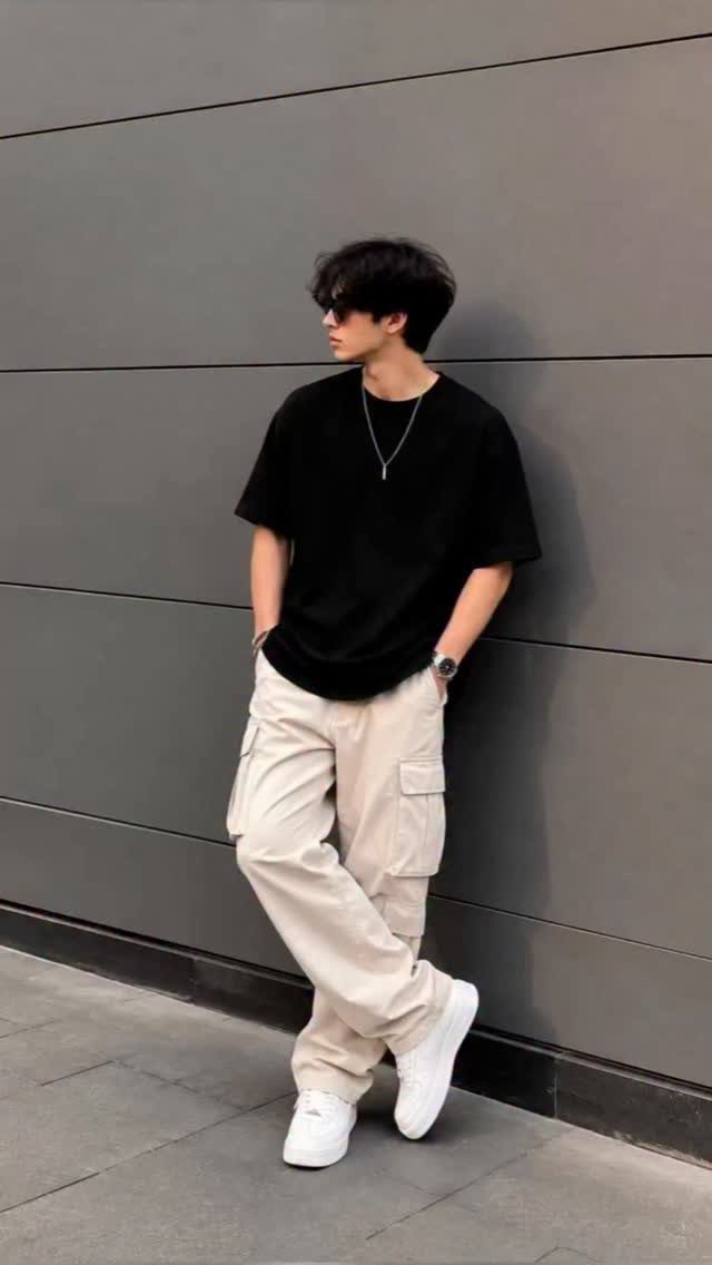 Man in black t-shirt and beige cargo pants leaning against a grey wall.