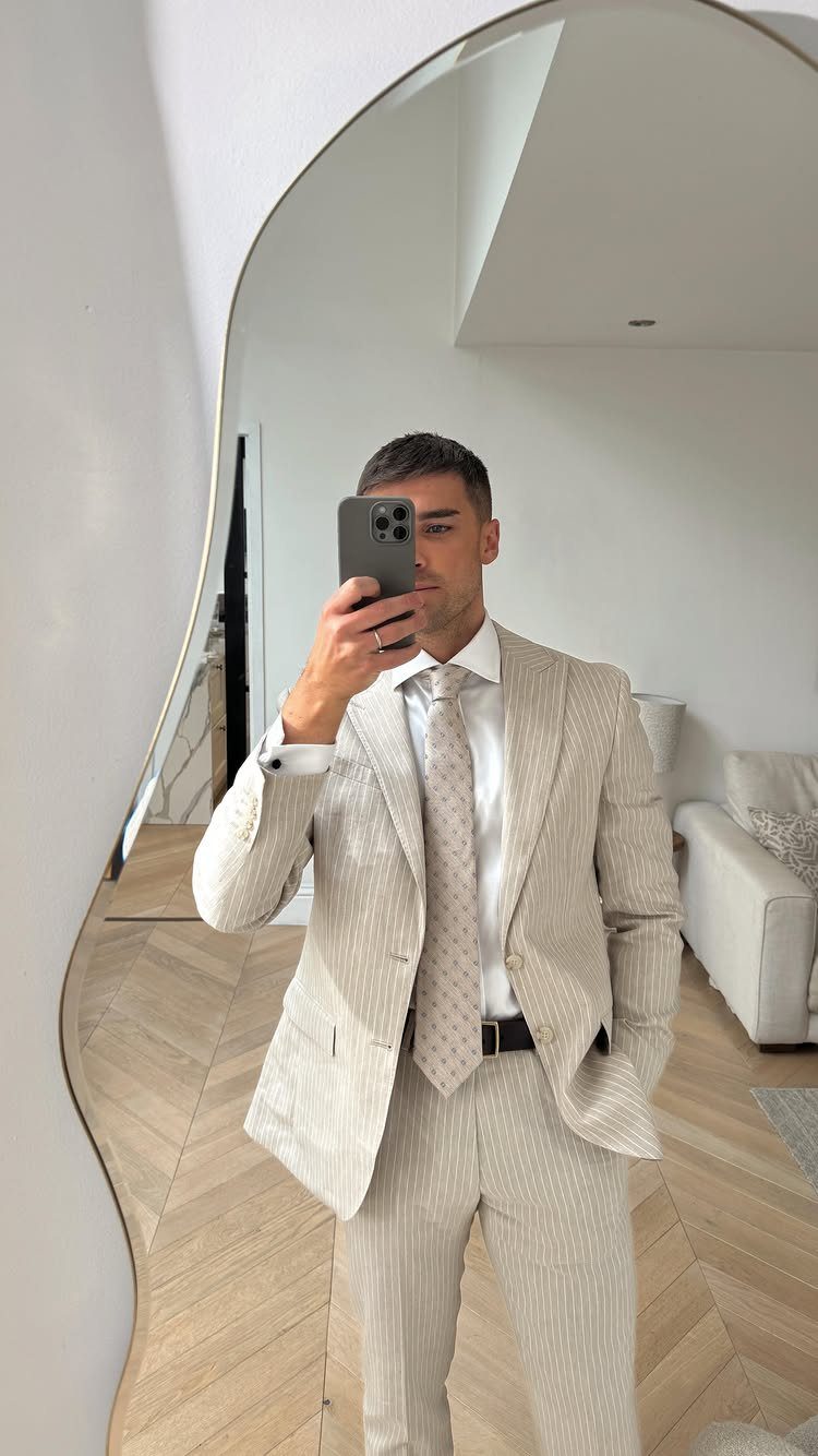 Man in a light beige textured suit and tie taking a mirror selfie.
