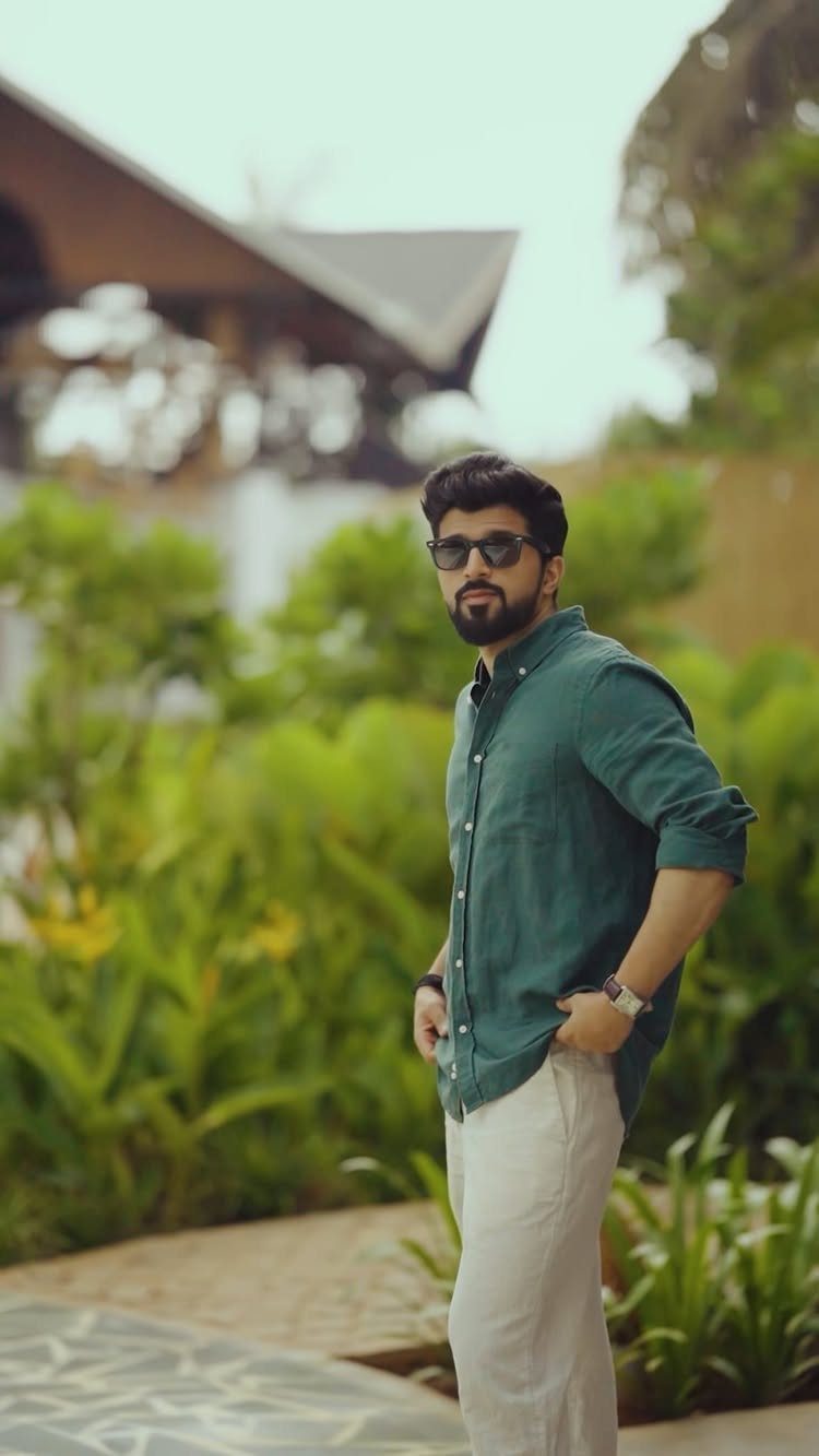 Man wearing a green button-down shirt and white trousers standing in a garden.