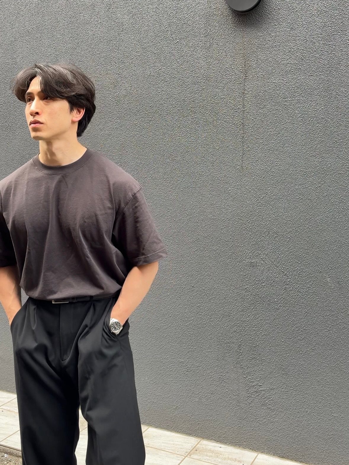 Man wearing a tucked dark grey t-shirt and black tailored trousers against a grey wall.