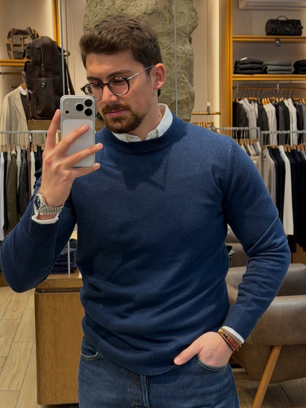 Man in a navy blue sweater and jeans taking a mirror selfie in store