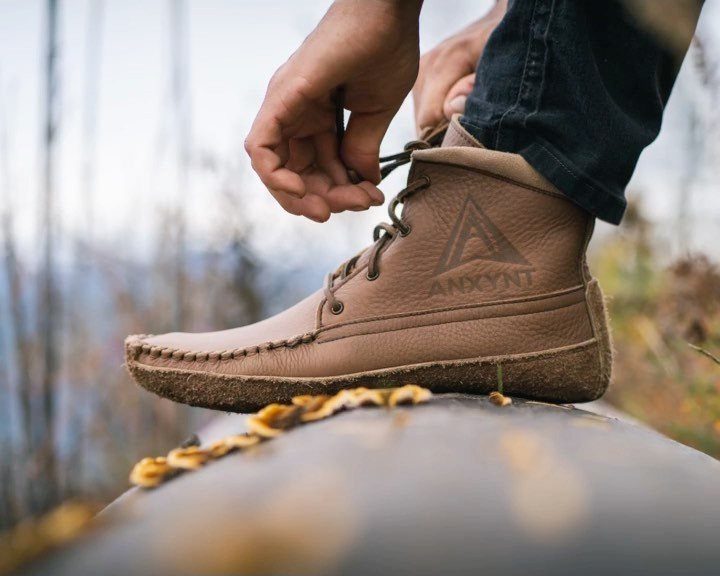 Brown leather moccasin boots being tied by a person wearing dark blue jeans.