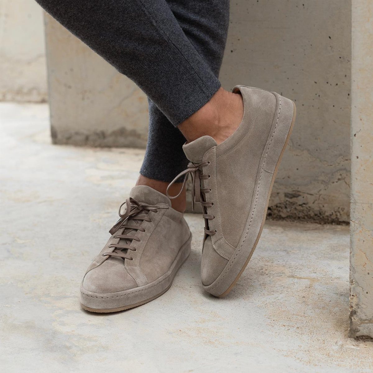 Beige suede sneakers worn with grey trousers on a concrete surface.