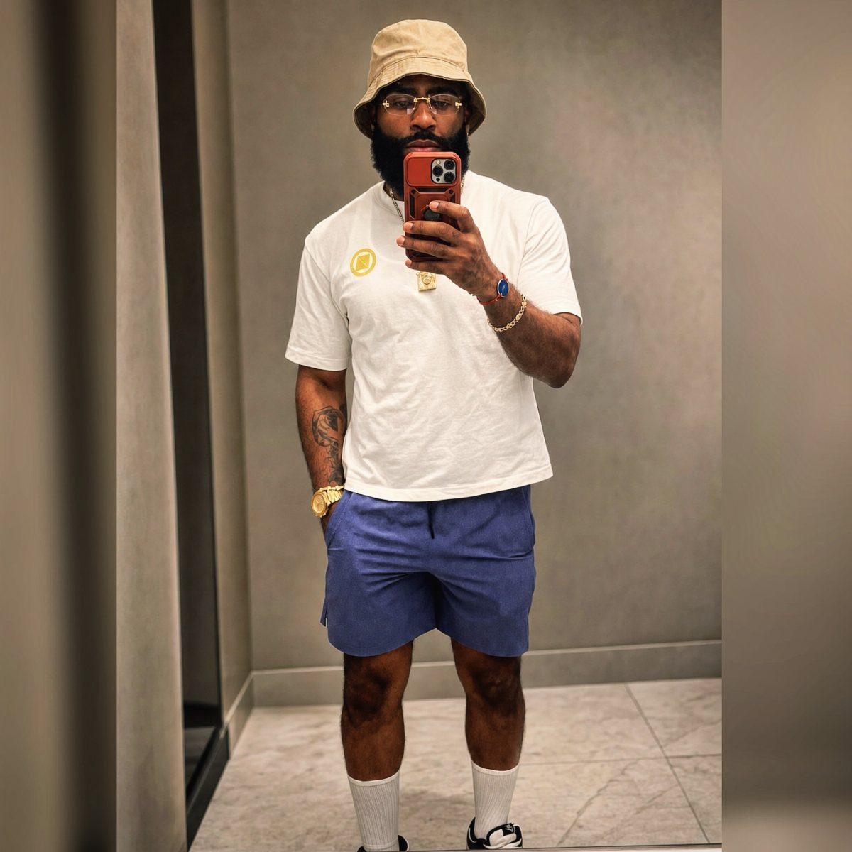 Man wearing a tan bucket hat, white shirt, and blue shorts in a mirror selfie.