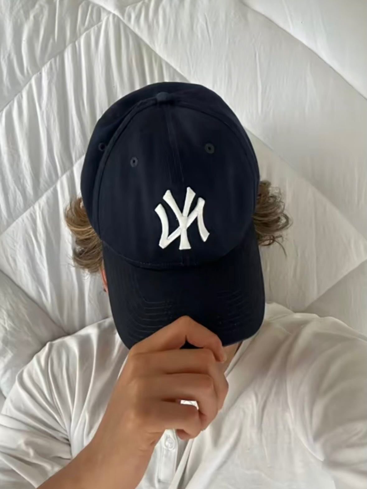 Person wearing a navy blue NY baseball cap and white t-shirt on white bedding.