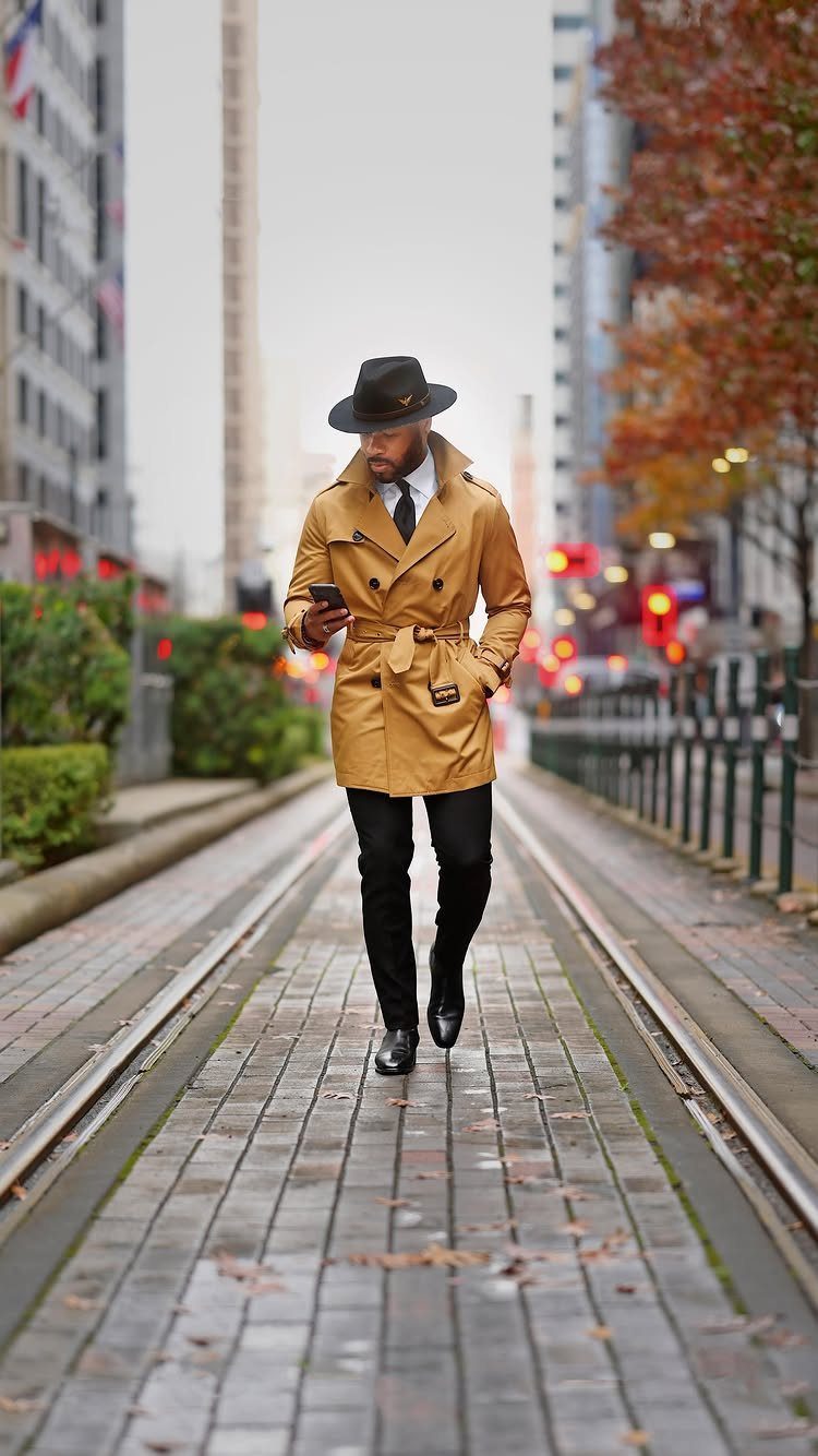 Man in black fedora and tan tailored trench coat standing on city street tracks.