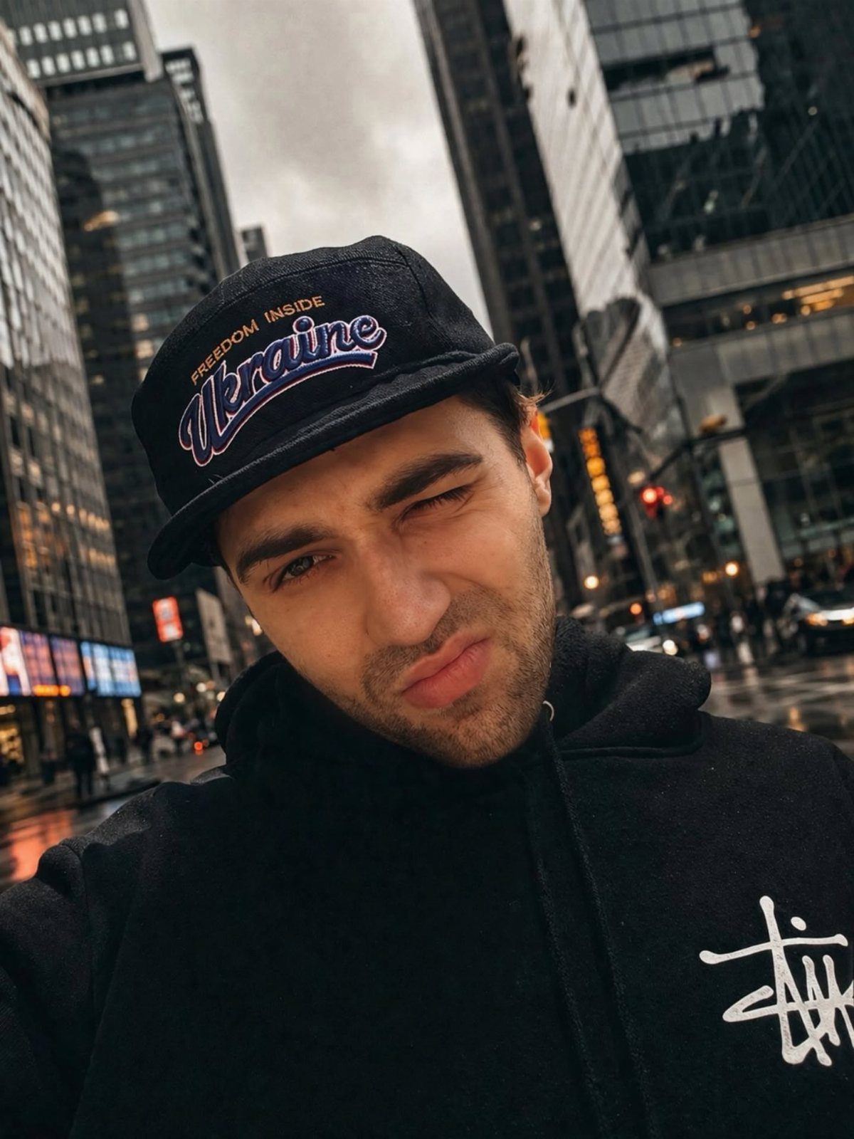 Man wearing a black snapback hat and black hoodie in a city street.