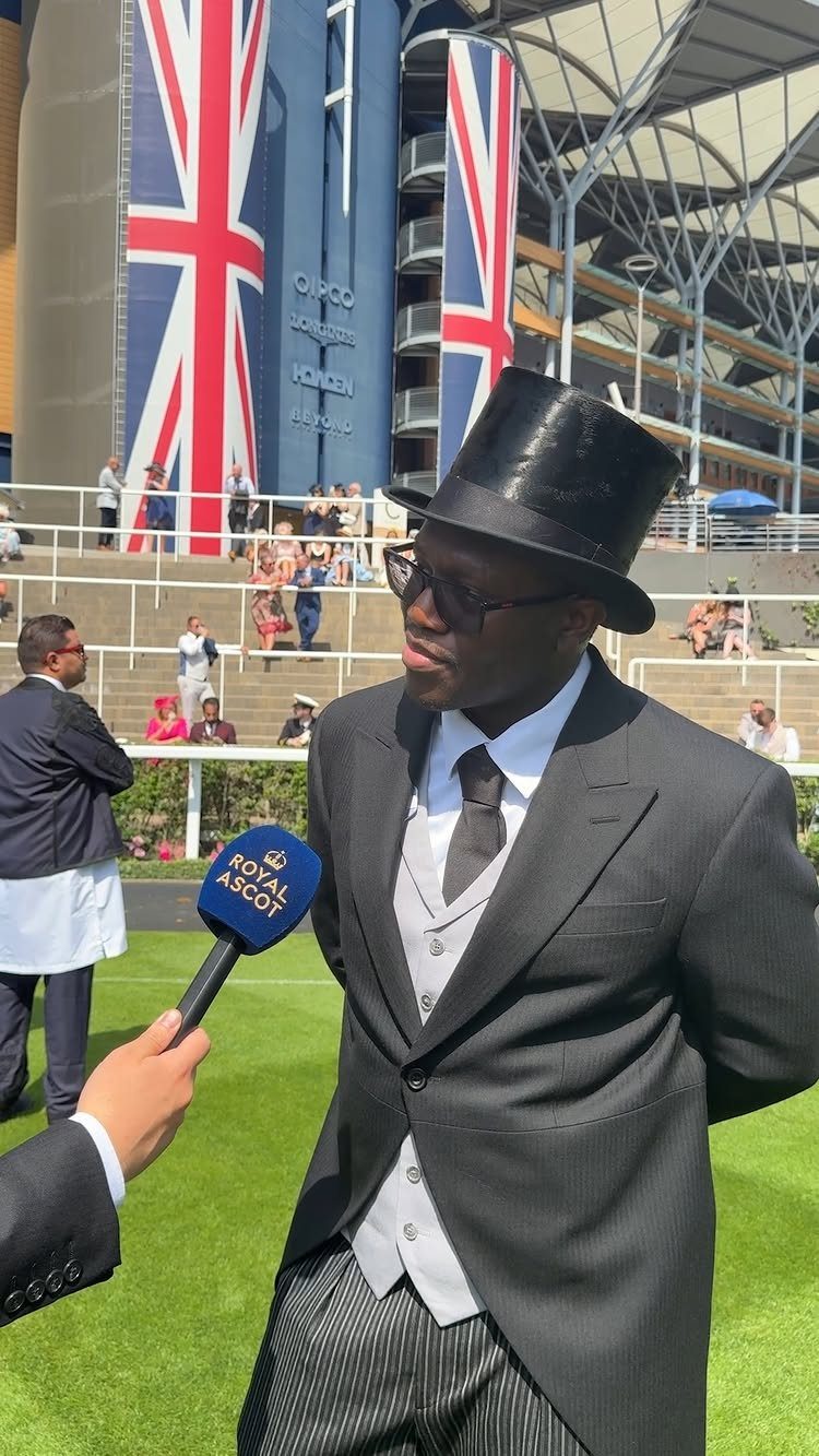 Man in a black top hat and formal morning suit being interviewed outdoors.