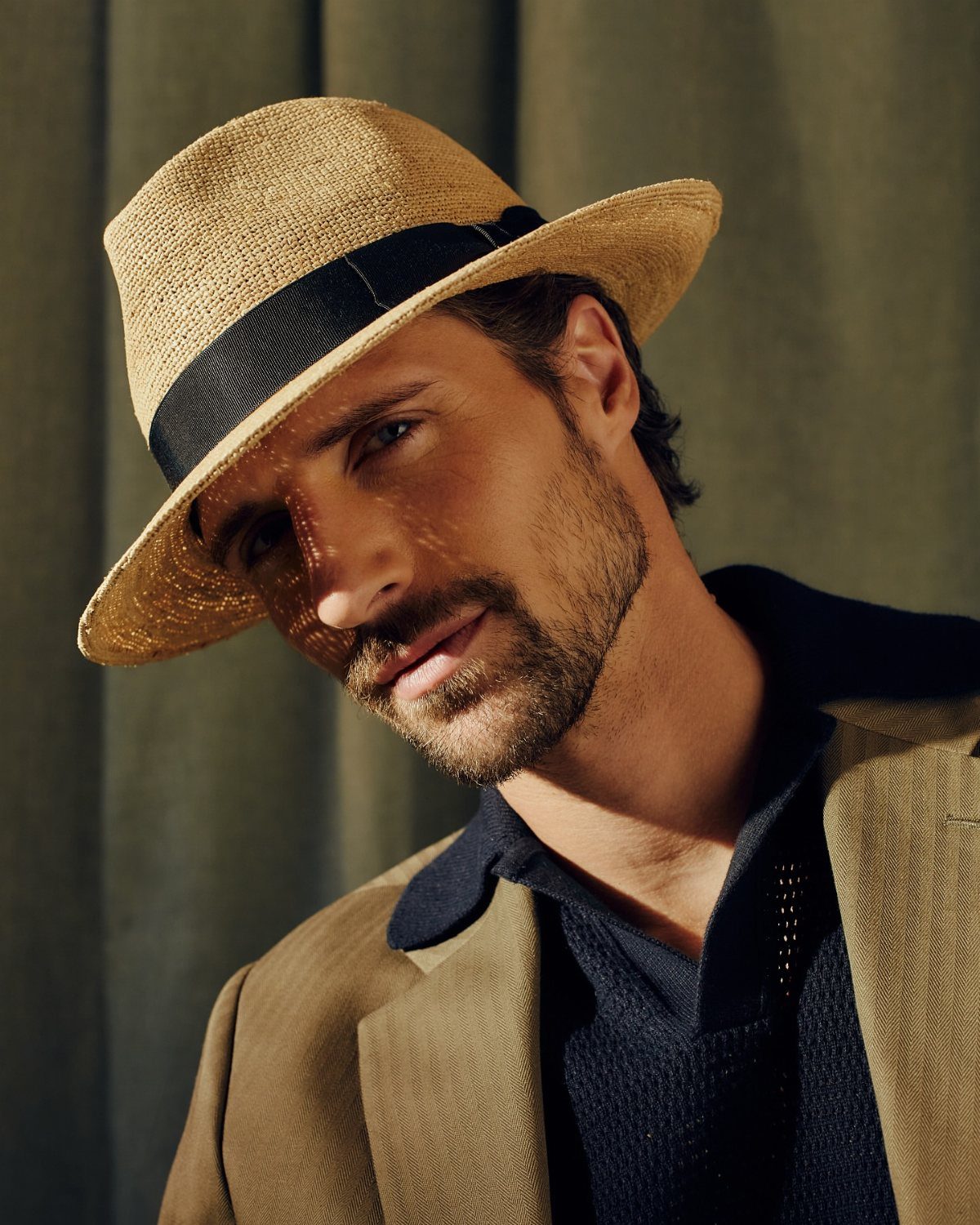Man wearing a straw trilby hat and tan blazer against a green backdrop.