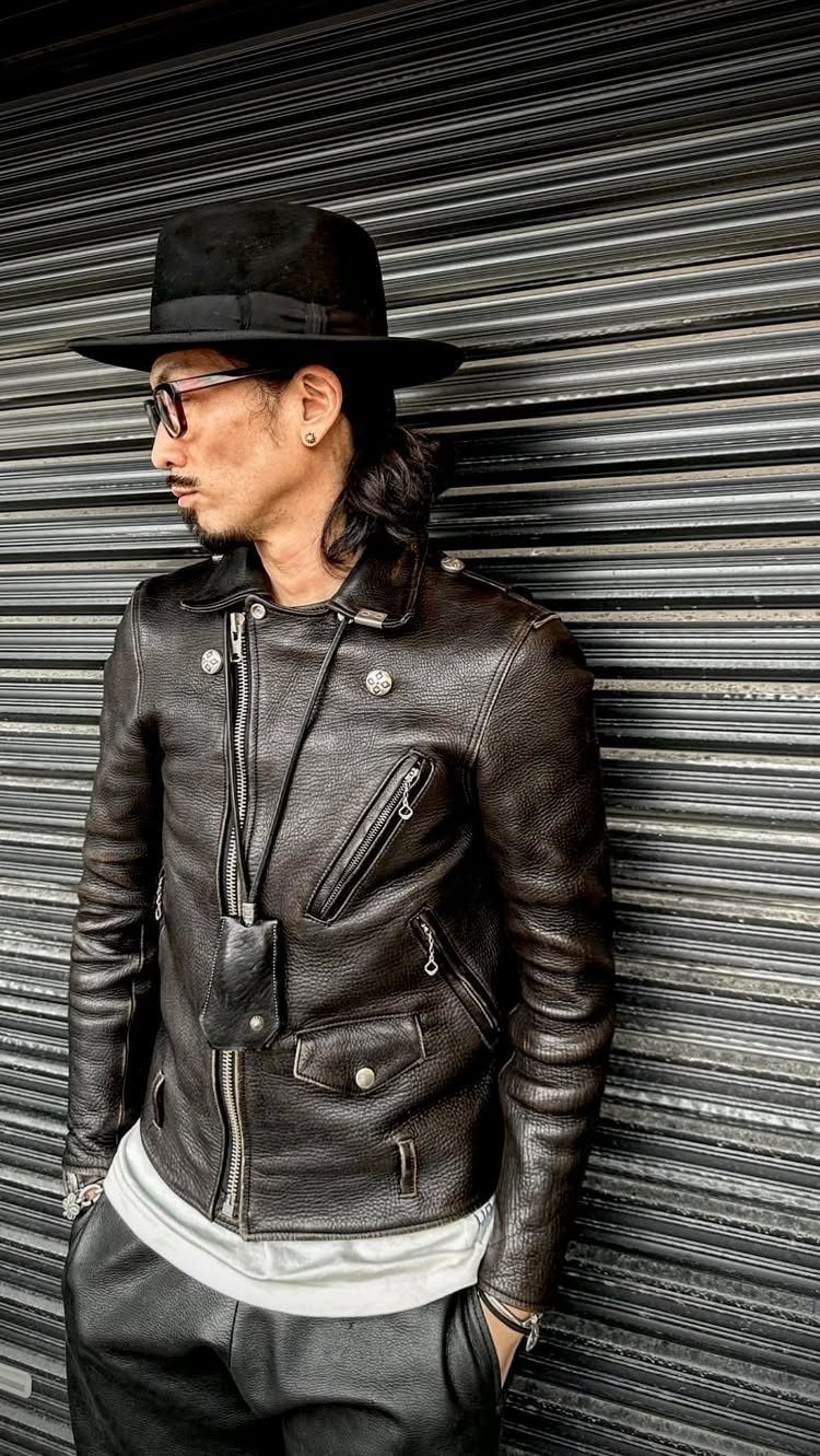 Man in black wide-brimmed hat and brown leather jacket posing by metal shutter.