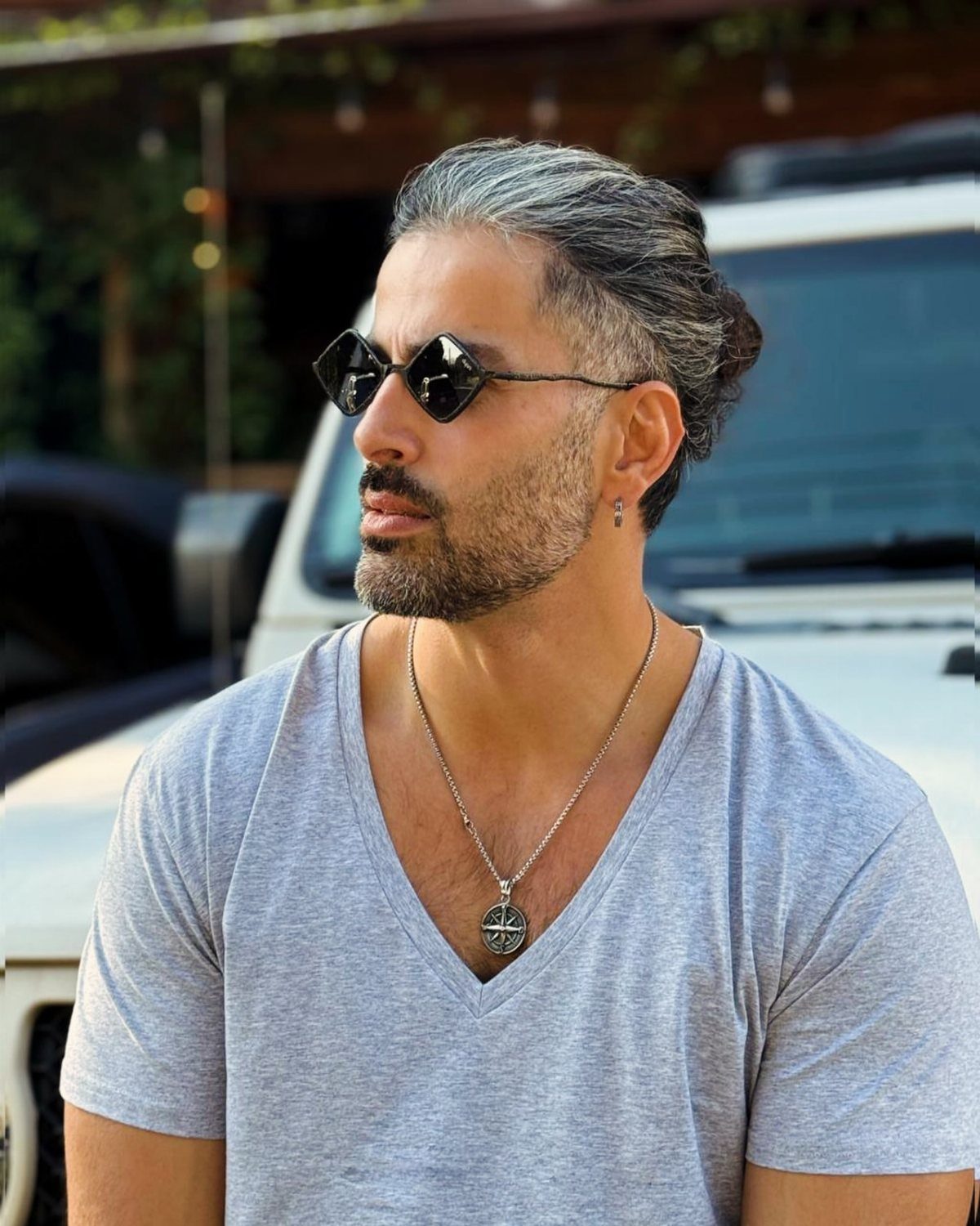Man with grey hair wearing black geometric sunglasses and a grey v-neck shirt.