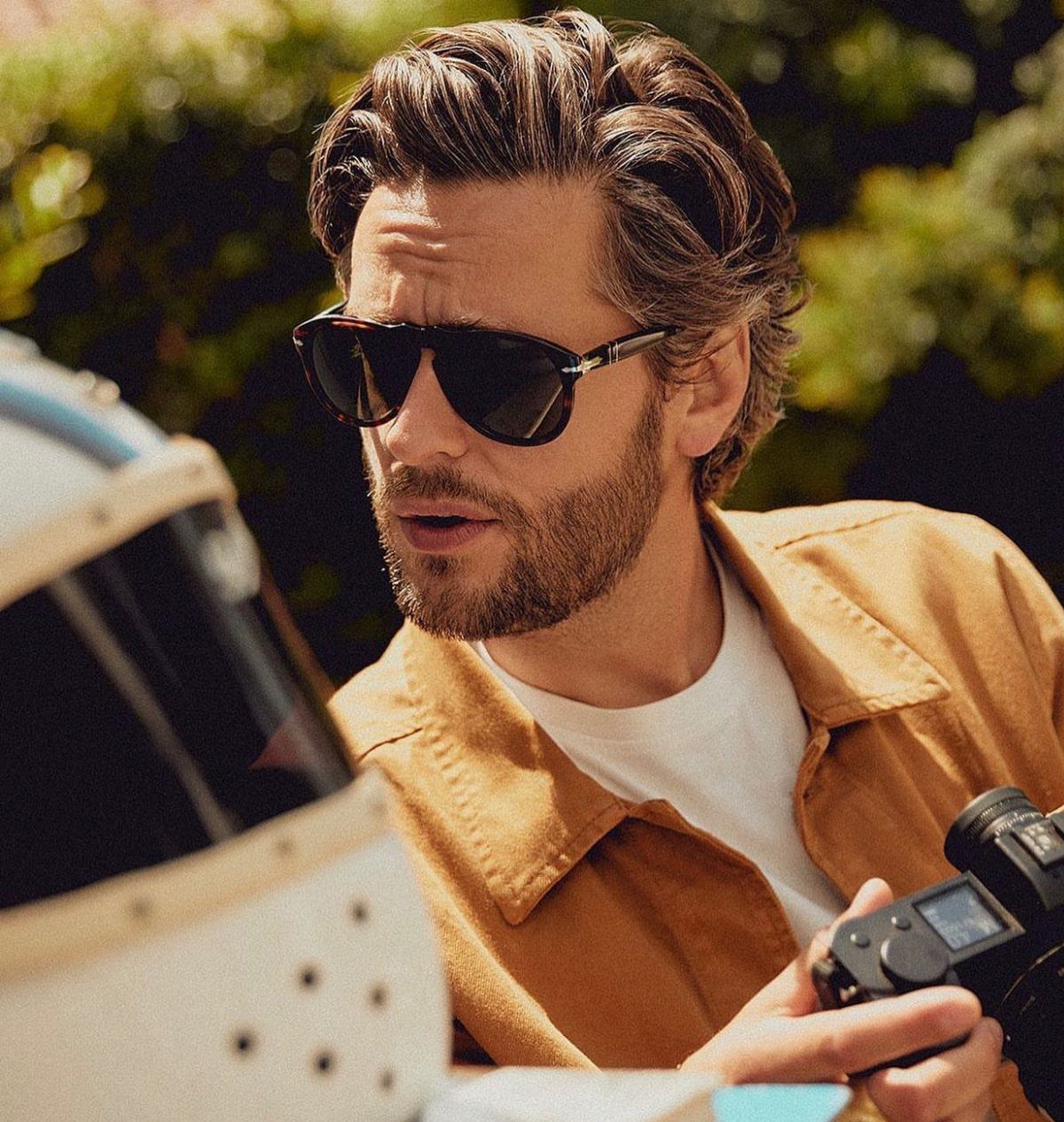 Man with beard wearing dark square-frame sunglasses and tan shirt holding a camera