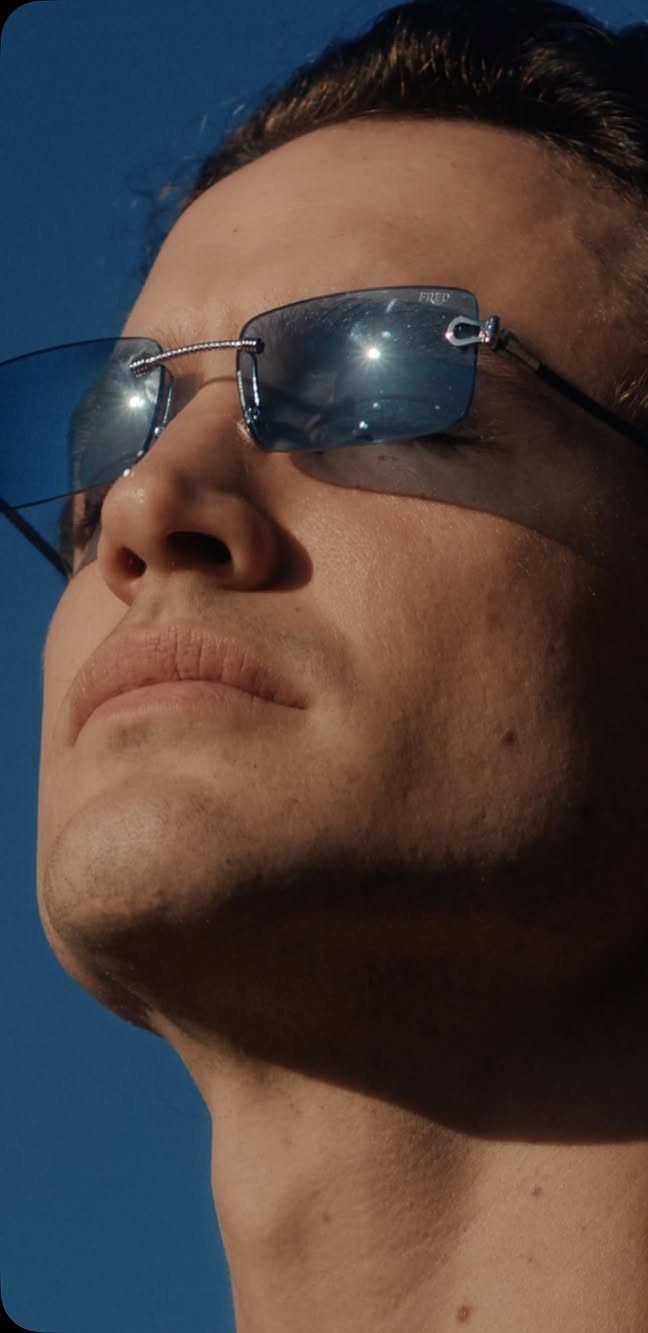 Man wearing rimless blue-tinted sunglasses looking upward against a clear blue sky.