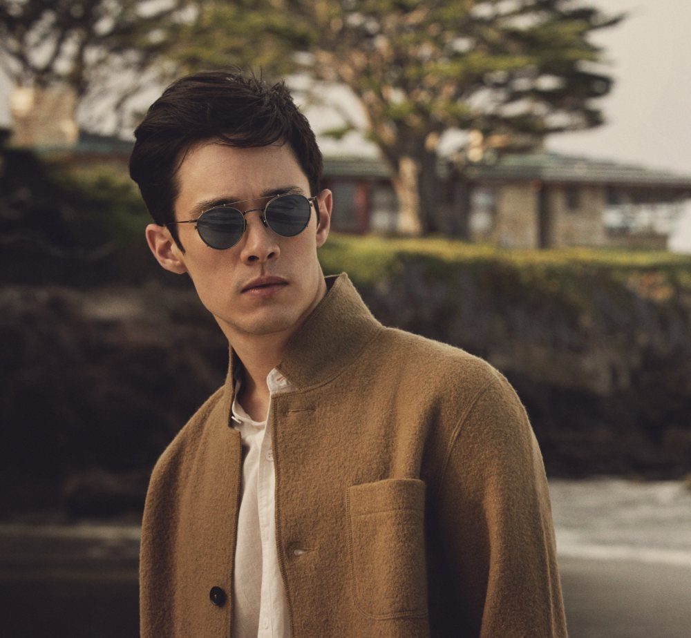 Man wearing round dark sunglasses and a brown wool jacket outdoors by the coast