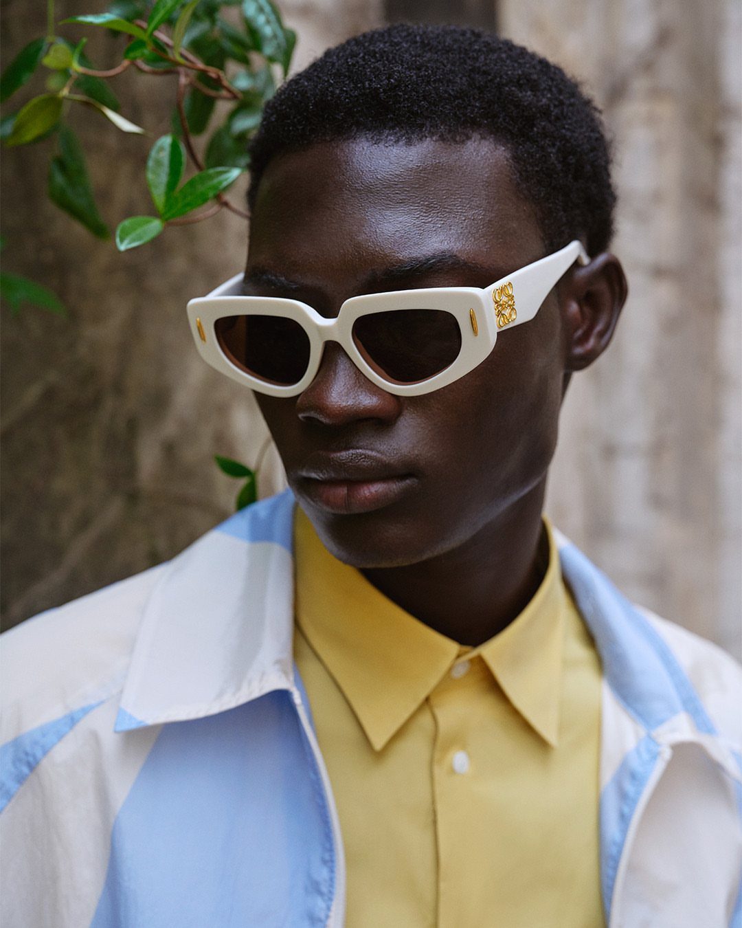 Man wearing thick white high fashion sunglasses, yellow shirt, and blue jacket.