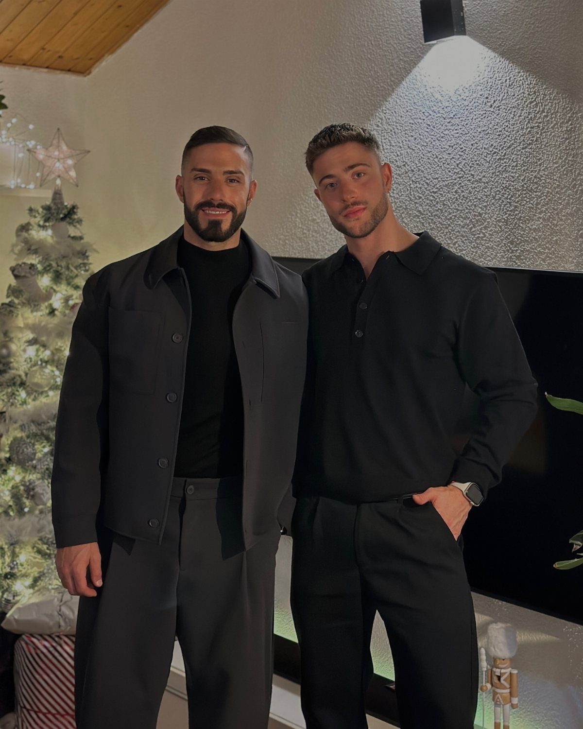 9 Men's Holiday Party Fashion Ideas That Actually Work 8 Two men in smart dark clothing posing beside a decorated Christmas tree indoors.