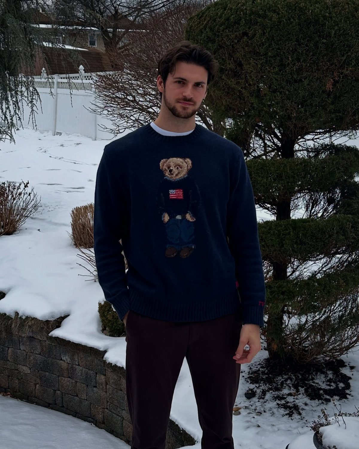 9 Men's Holiday Party Fashion Ideas That Actually Work 5 Man wearing a navy blue bear sweater and brown pants in a snowy yard.