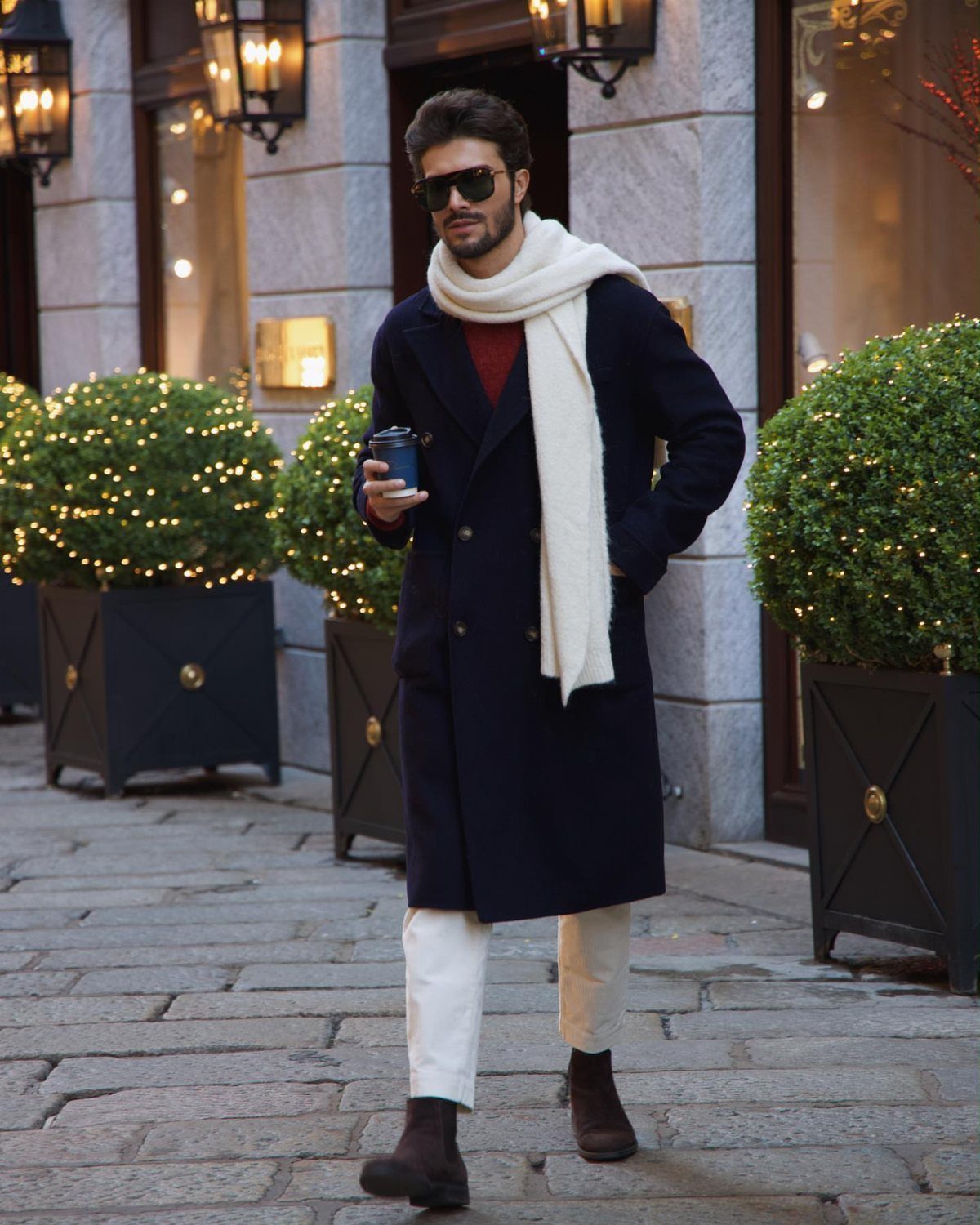 9 Men's Holiday Party Fashion Ideas That Actually Work 6 Man in navy overcoat and white scarf standing on a festive cobblestone street