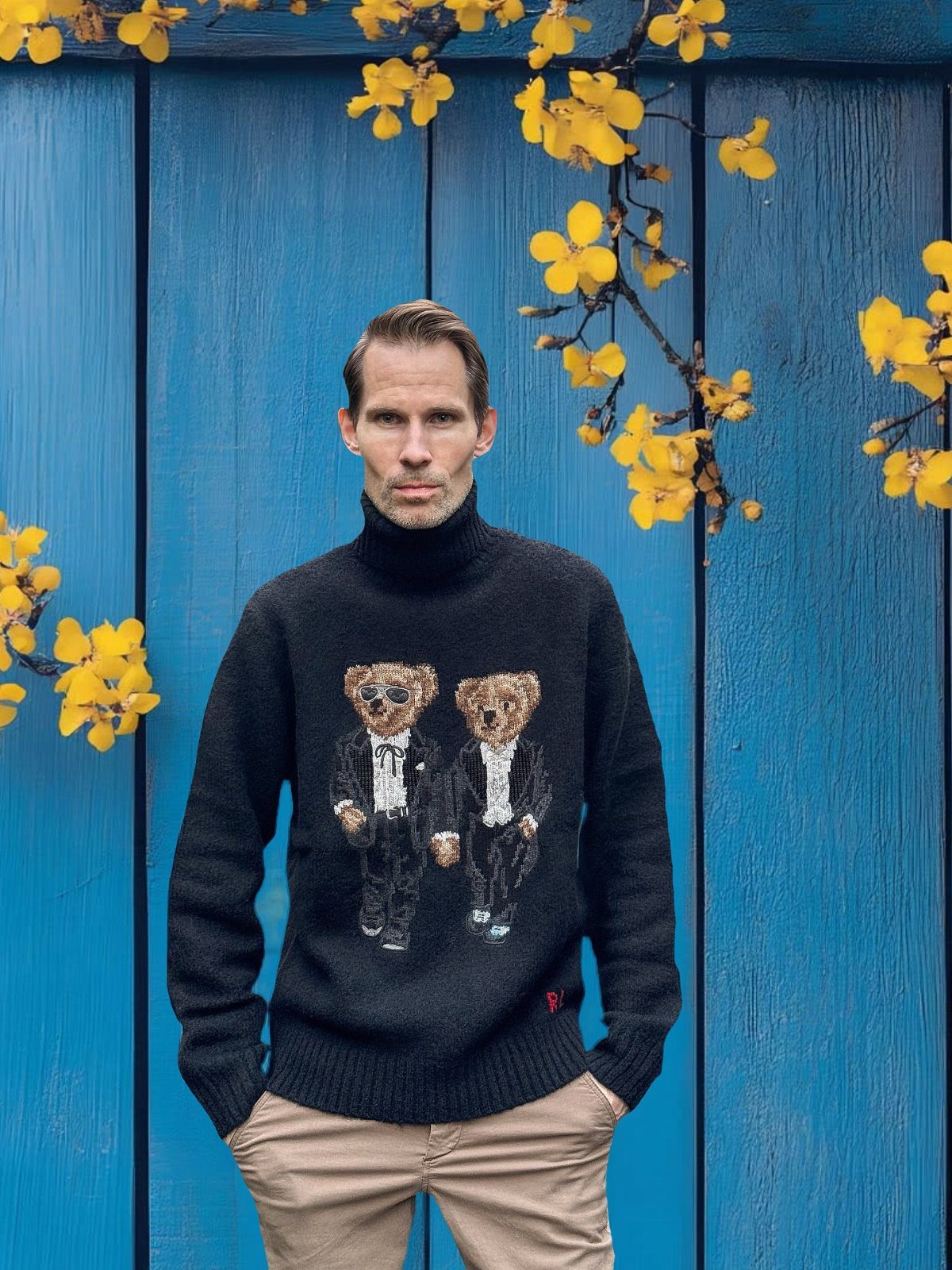 9 Men's Holiday Party Fashion Ideas That Actually Work 9 Man in black bear-themed turtleneck sweater and tan chinos against blue wooden wall.