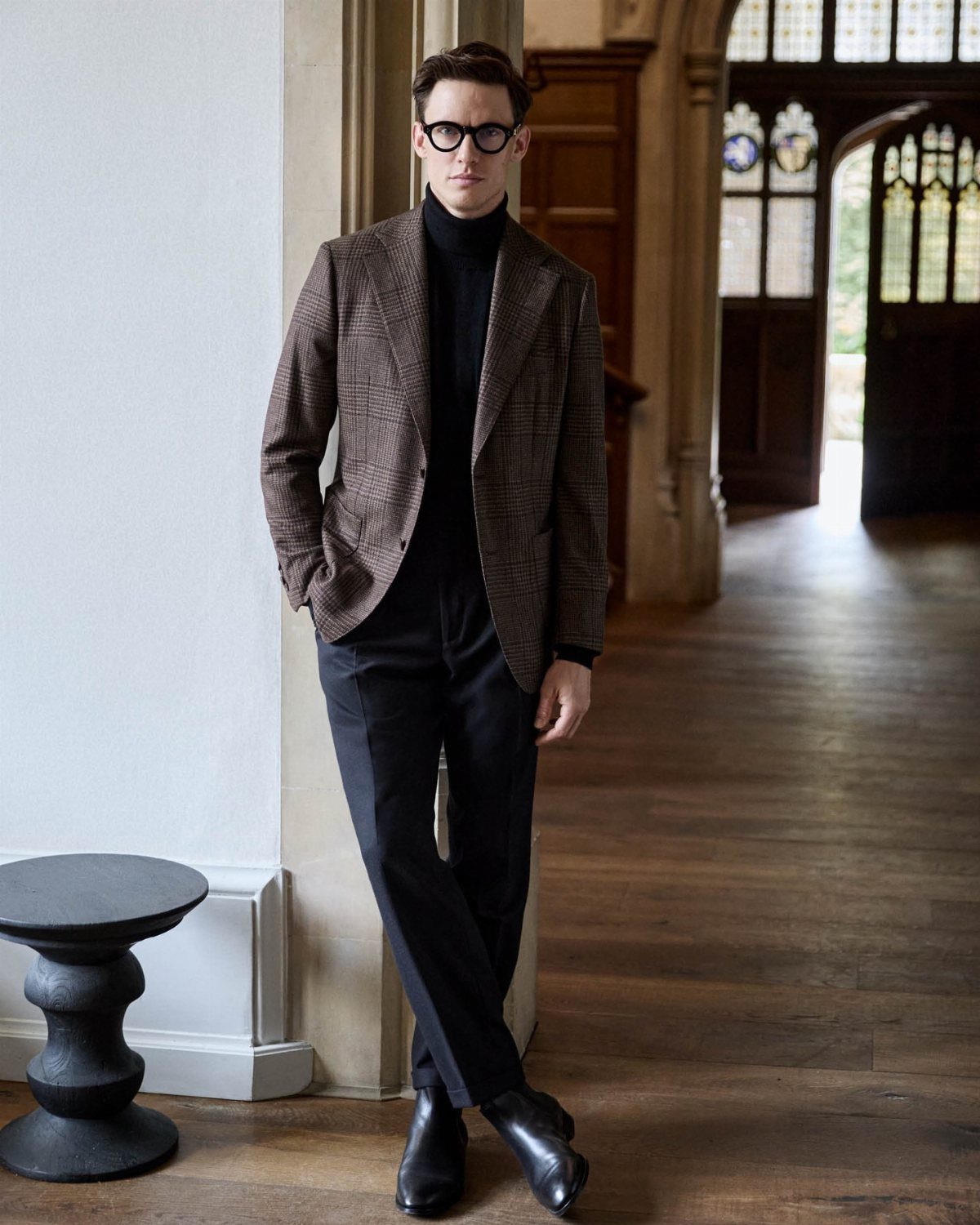 9 Men's Holiday Party Fashion Ideas That Actually Work 1 Man in brown patterned blazer and black turtleneck posing in a wooden hallway.