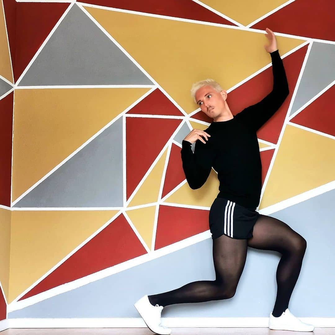 Man in black shirt, athletic shorts, and black pantyhose posing by geometric wall.
