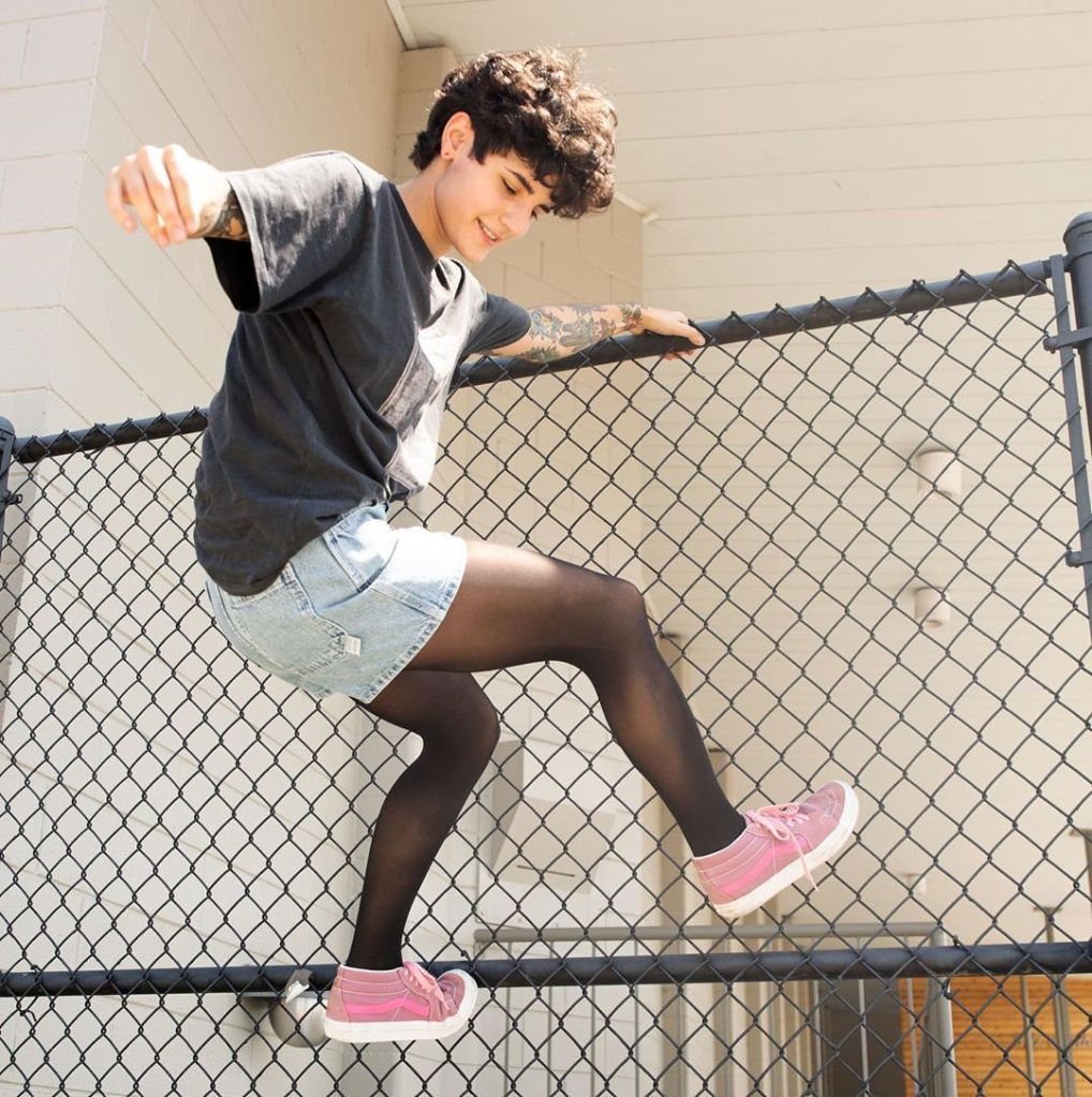Young man wearing denim shorts and black pantyhose posing by a chain-link fence.