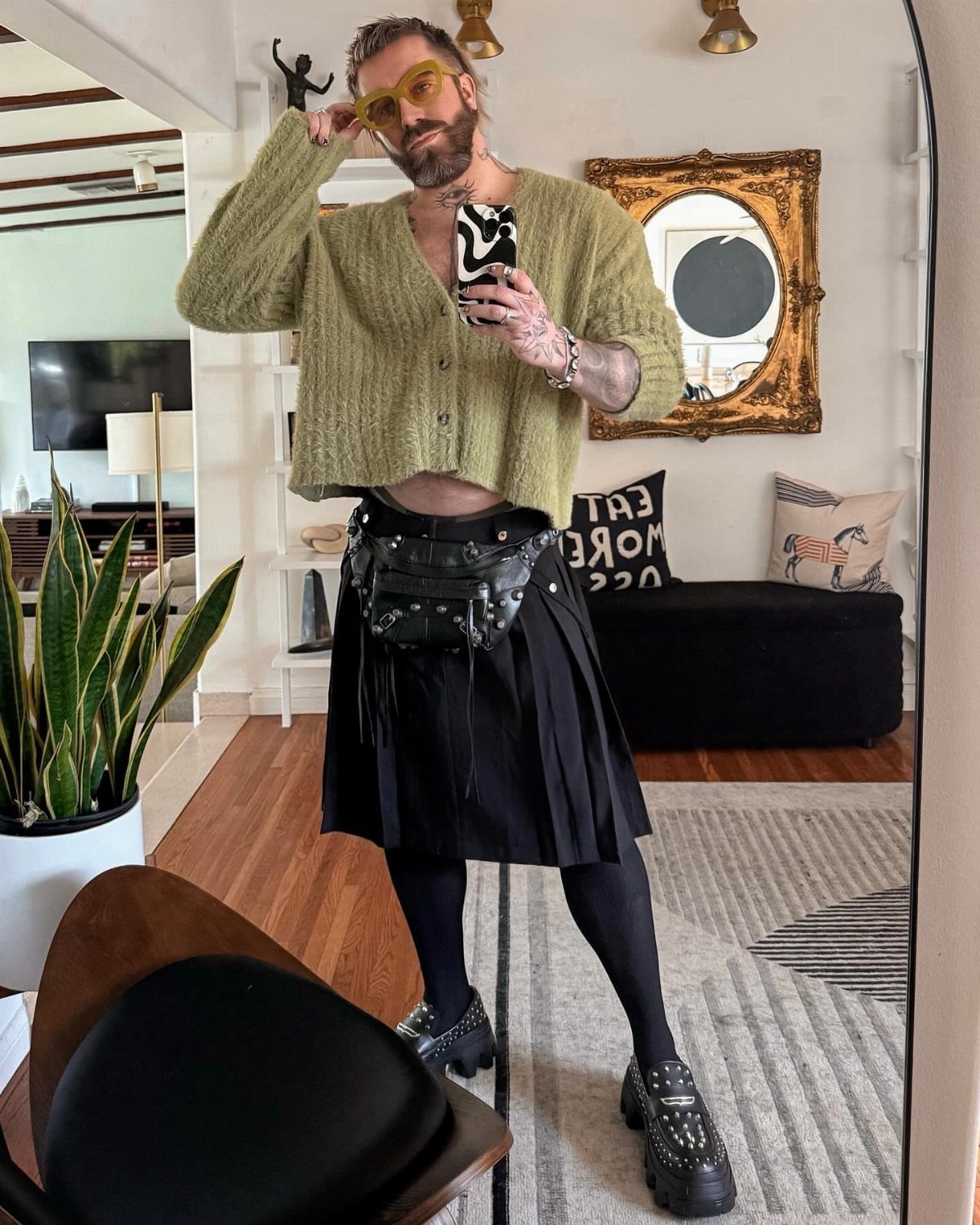 Man wearing a black pleated mini kilt, black tights, and green cardigan.