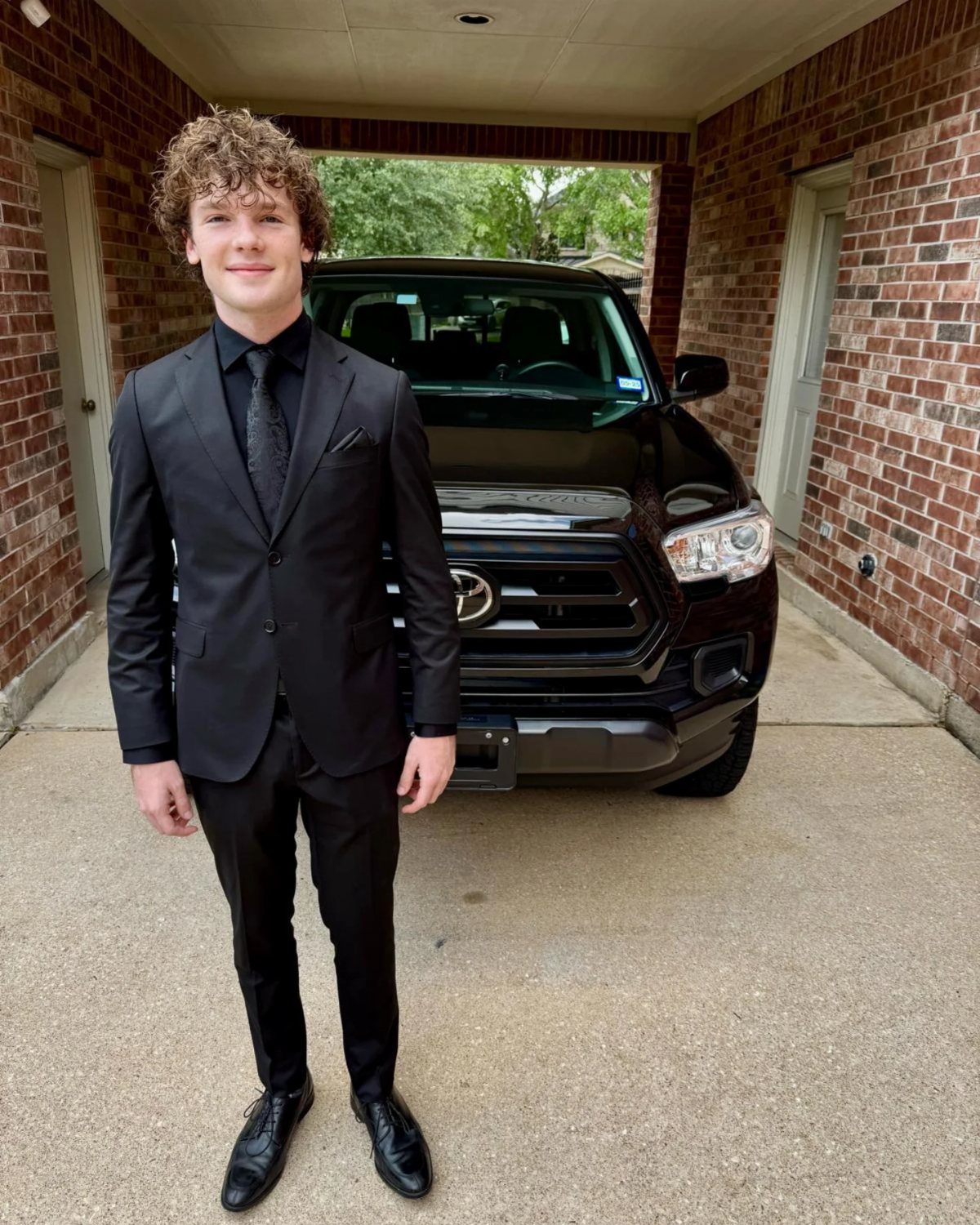 Young man in a formal black suit and tie standing before a black truck.