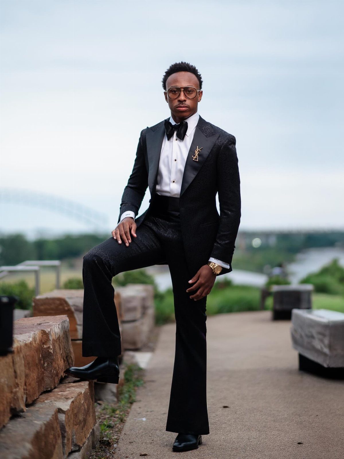 Young man in a black tuxedo and bow tie posing outdoors by a stone wall.
