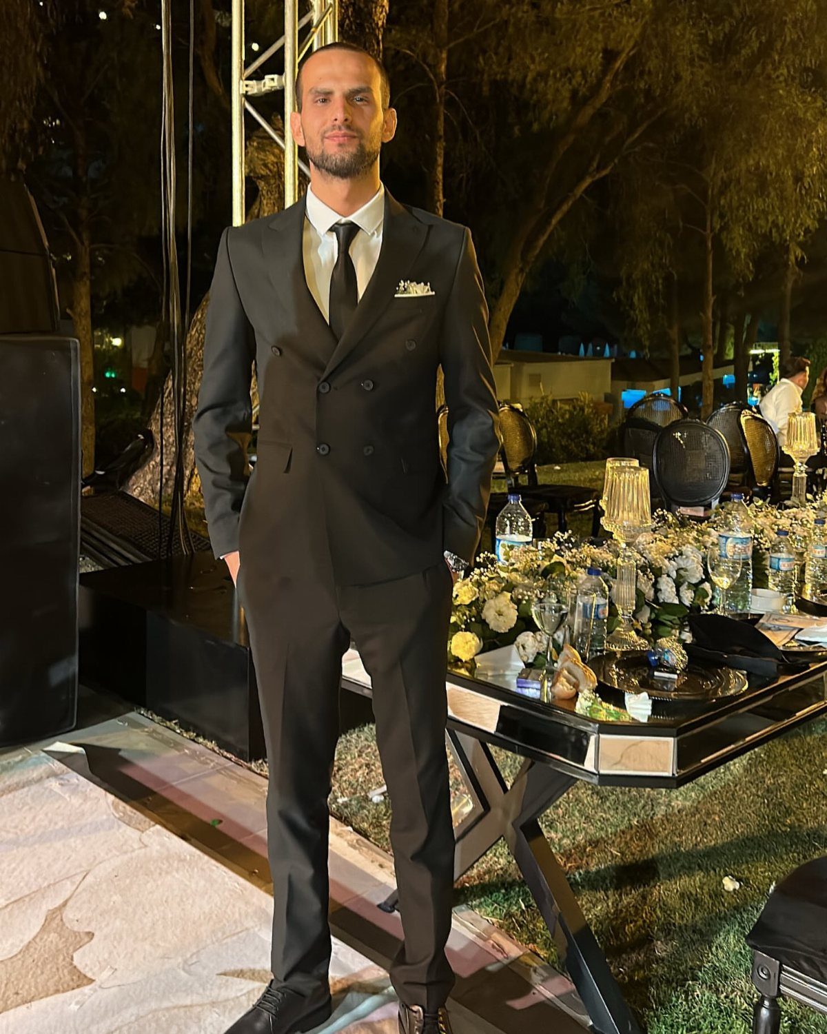 Man in a black double-breasted suit and tie posing at an outdoor event.