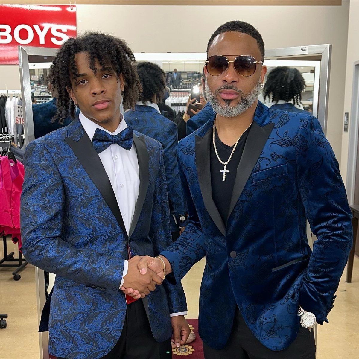 Two men shaking hands wearing ornate blue patterned tuxedo jackets in a store
