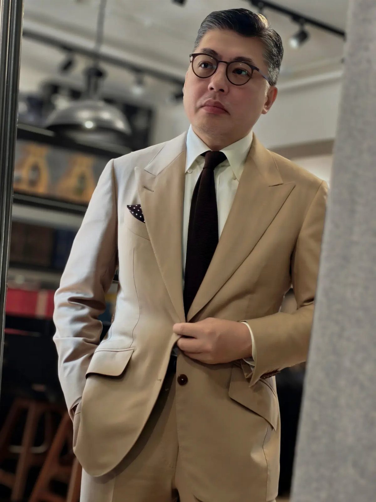 Man in a tan suit with a brown tie and glasses posing indoors.