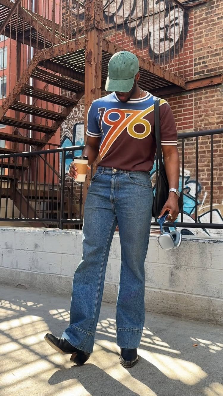 Man in vintage brown t-shirt, green cap, and wide-leg jeans standing outdoors.