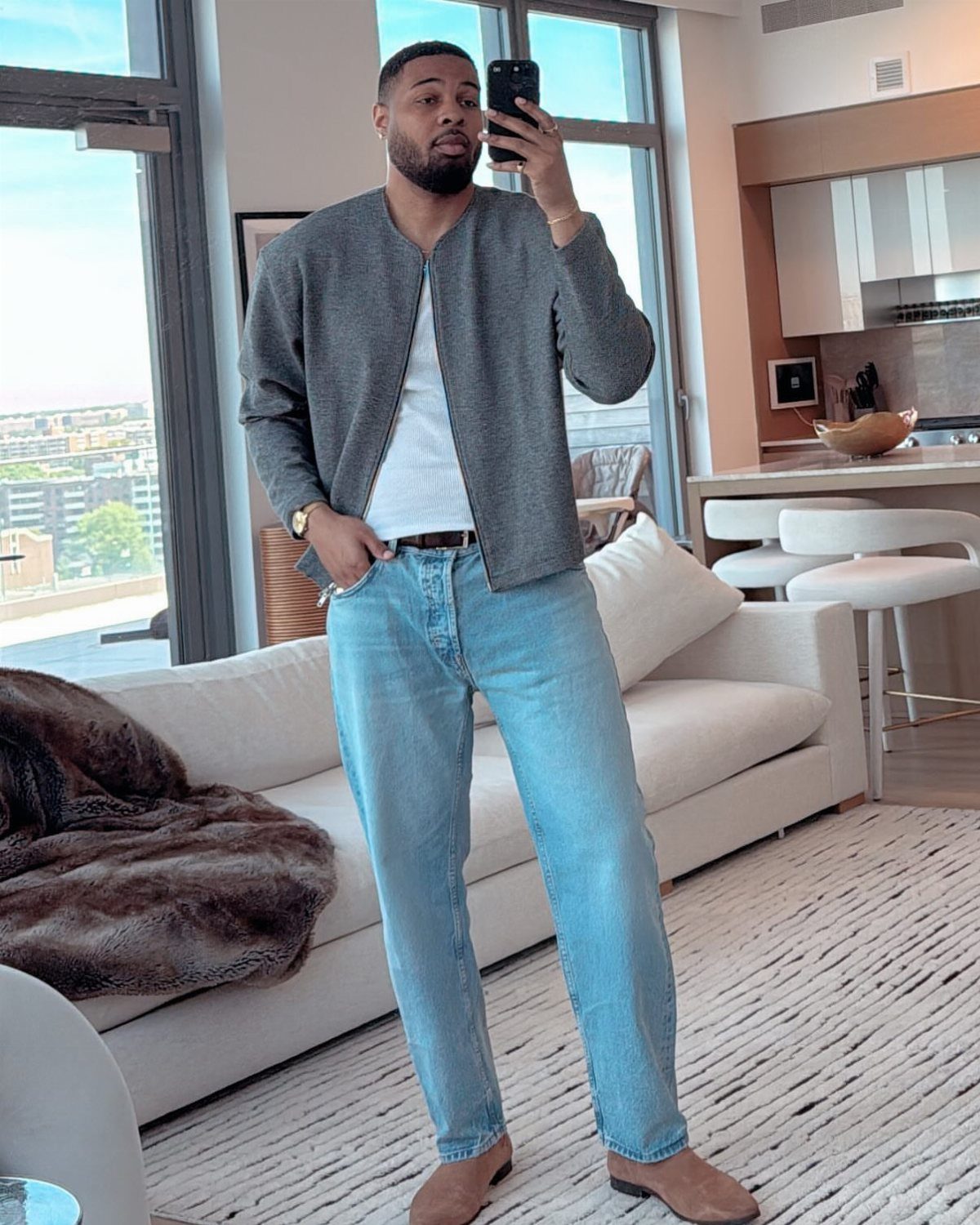 Man wearing a grey jacket and light blue straight-leg jeans in a modern room.