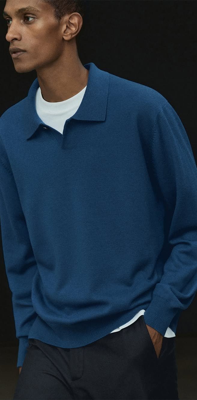 Man wearing a blue long-sleeved polo shirt and dark trousers against a dark background.