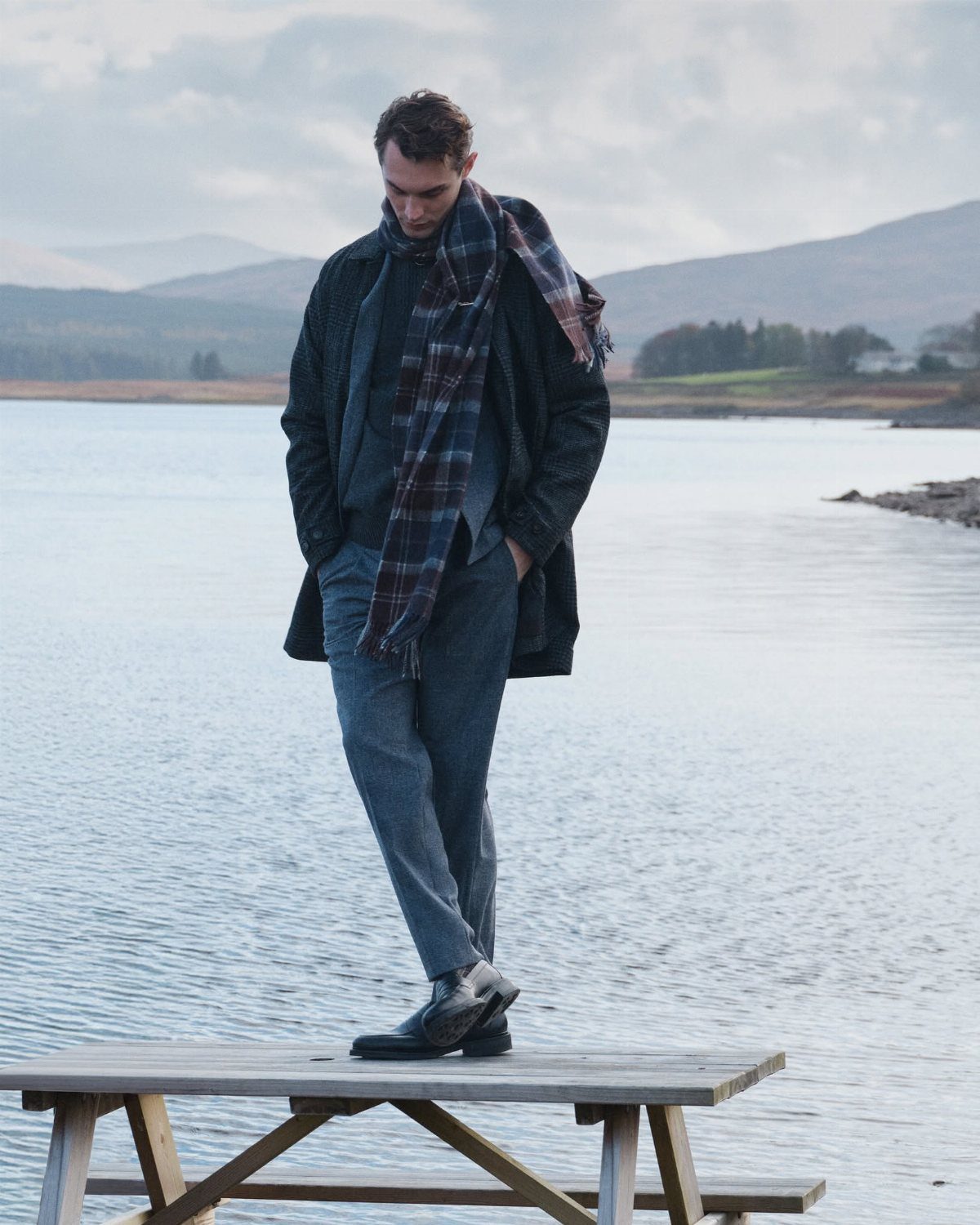 Man wearing a dark jacket, plaid scarf, and grey trousers standing by a lake.