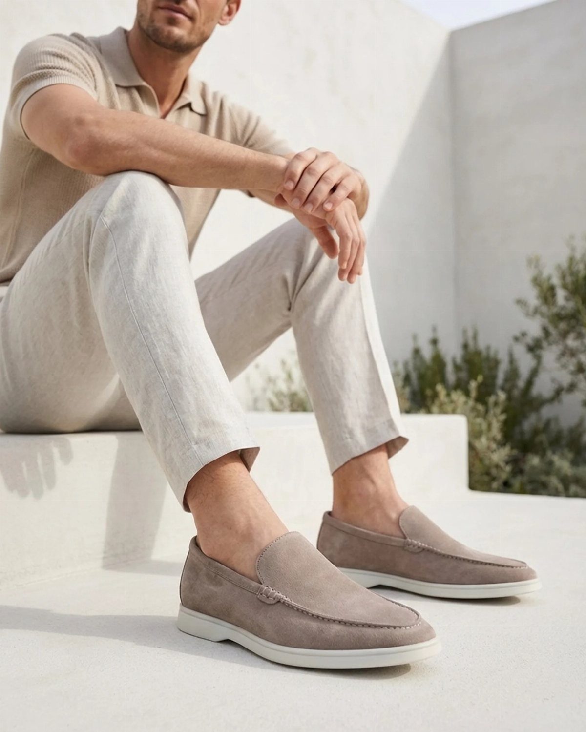 Man wearing grey suede loafers and light linen trousers sitting on a white ledge.
