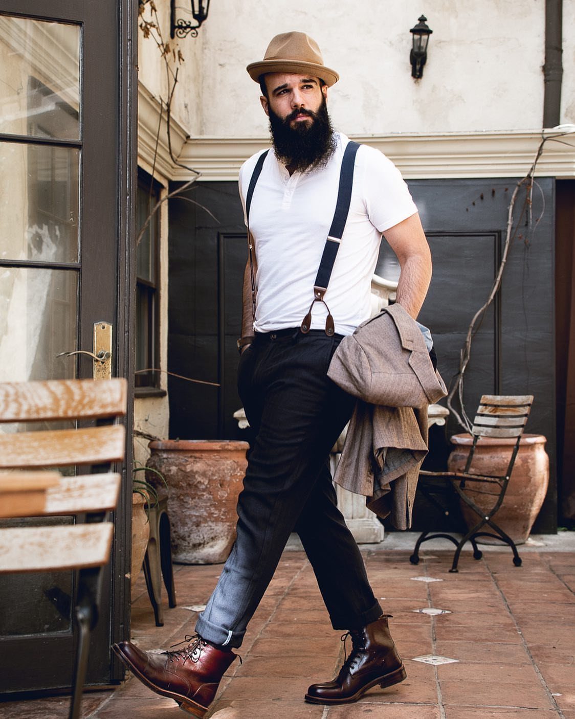 Bearded man in a white t-shirt and dark blue suspenders walking on a patio.