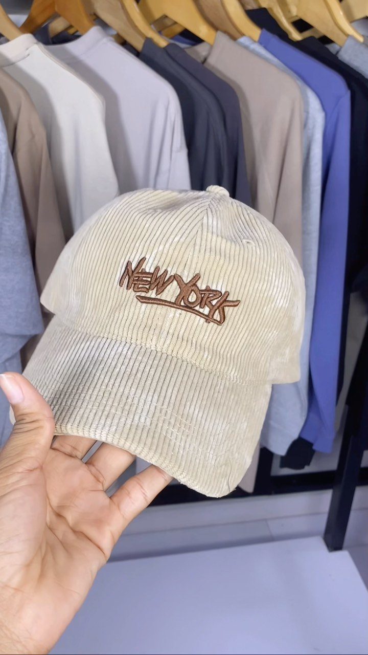 Beige corduroy baseball cap with New York embroidery held against clothing rack